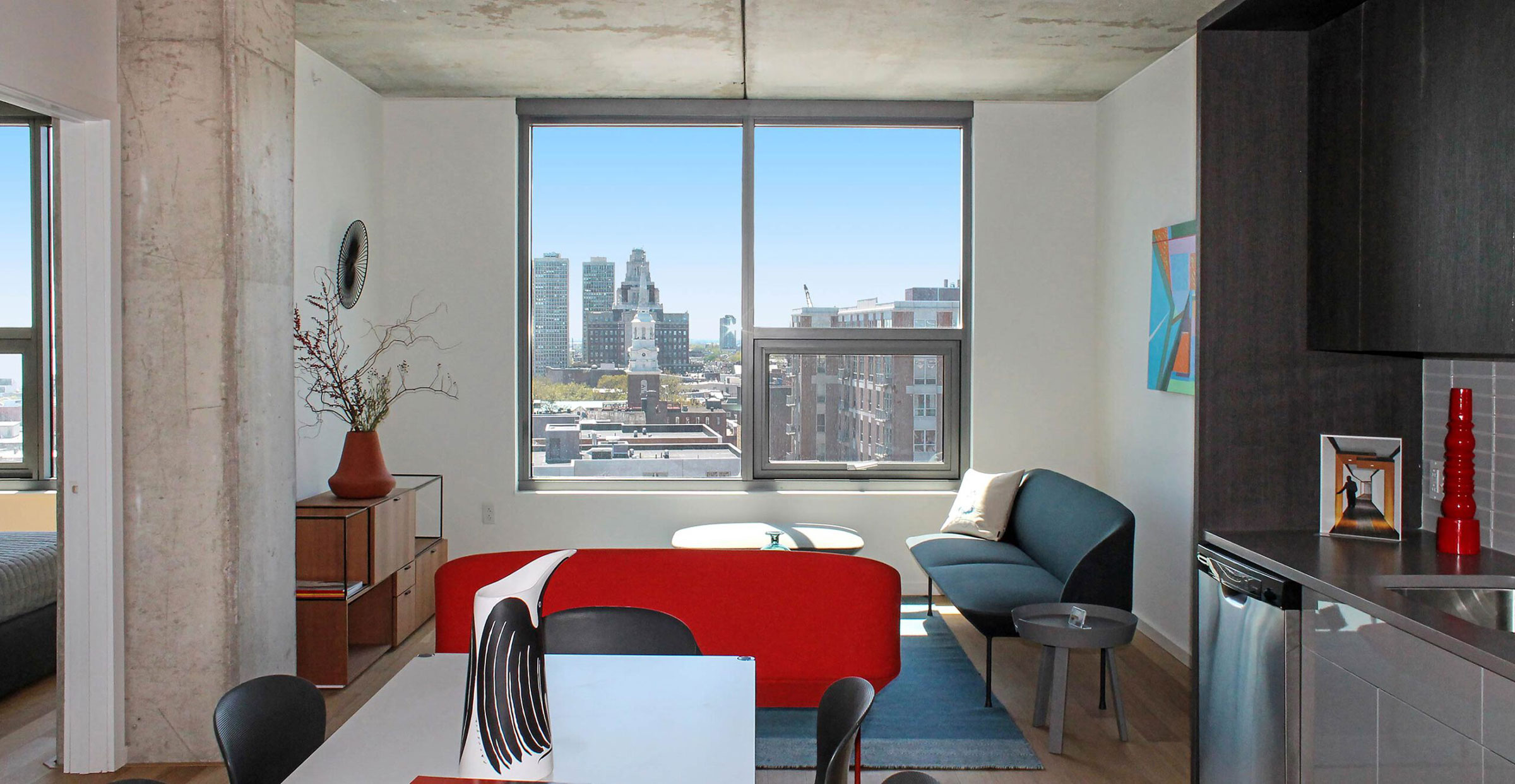 Modern apartment living room with large window view of cityscape. Features red sofa, blue chair, vase with branches, and abstract wall art. Bright and airy ambiance.