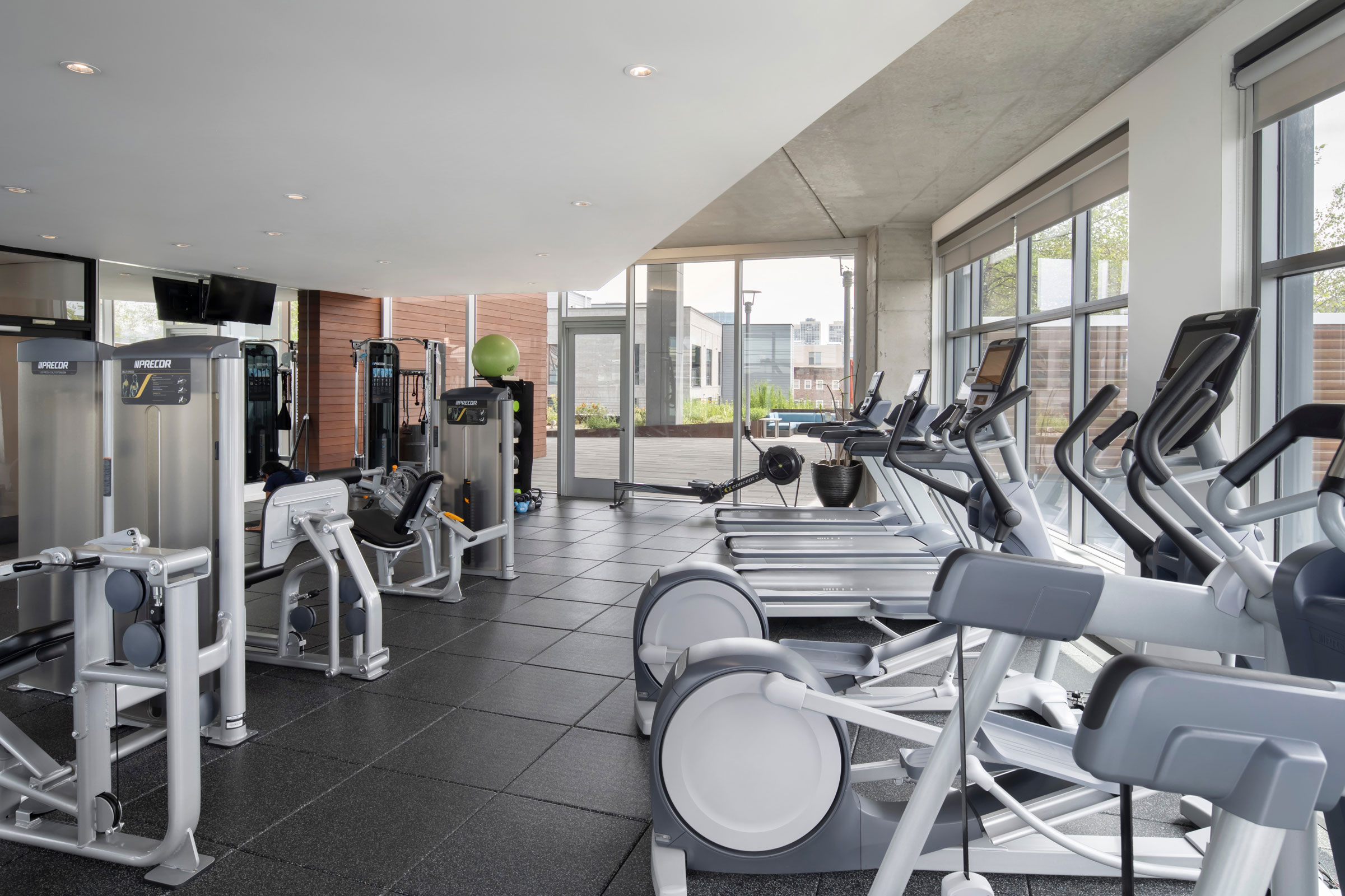 Modern gym interior with large windows, featuring treadmills, elliptical machines, and weight equipment. Bright lighting creates an inviting atmosphere.