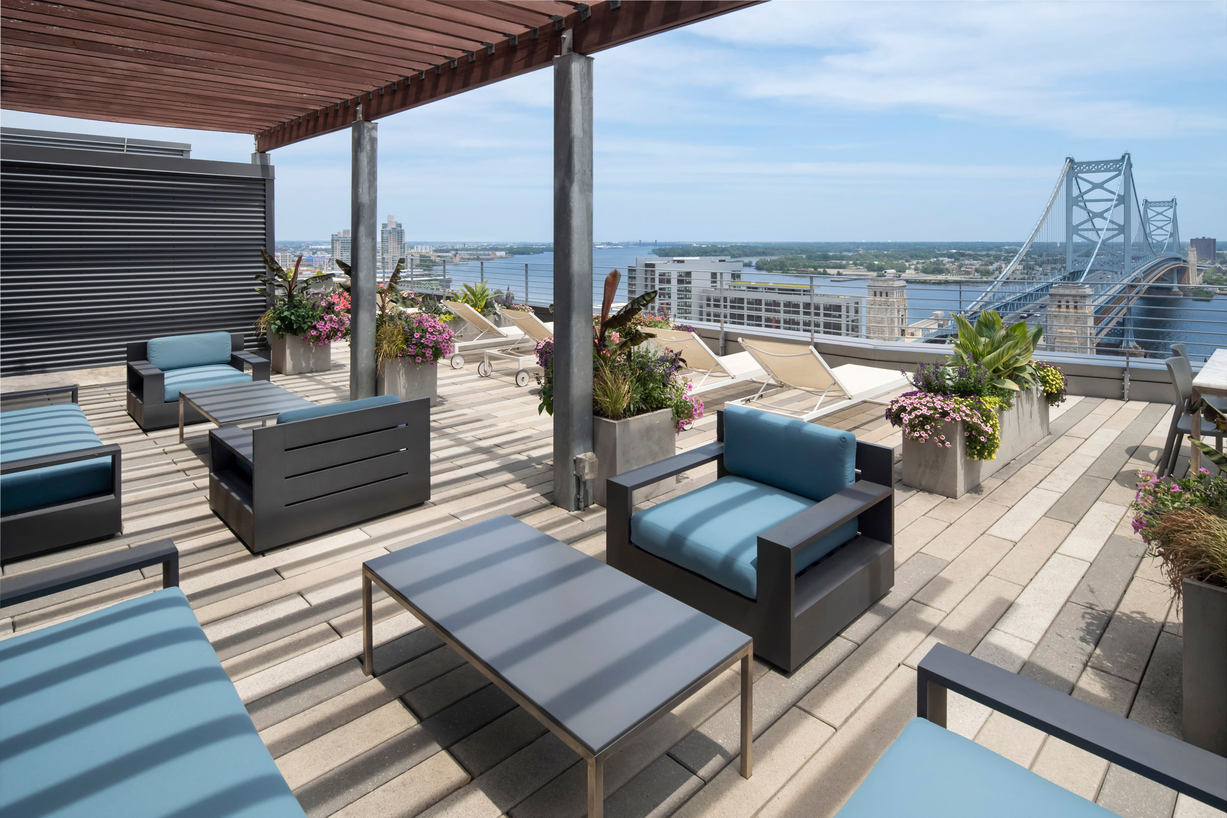 Rooftop terrace with modern seating and blue cushions under a wooden pergola. Lush potted plants and lounge chairs overlook a bridge and river view.