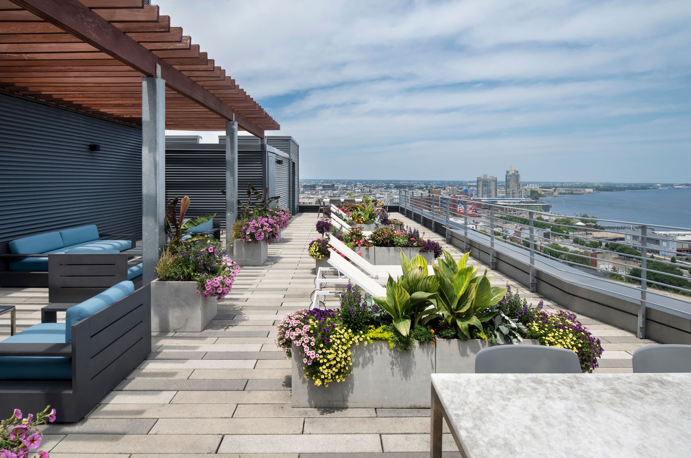 Rooftop terrace with modern seating, colorful flower planters, and a wooden pergola. Overlooks cityscape and river under a partly cloudy sky. Relaxing ambiance.