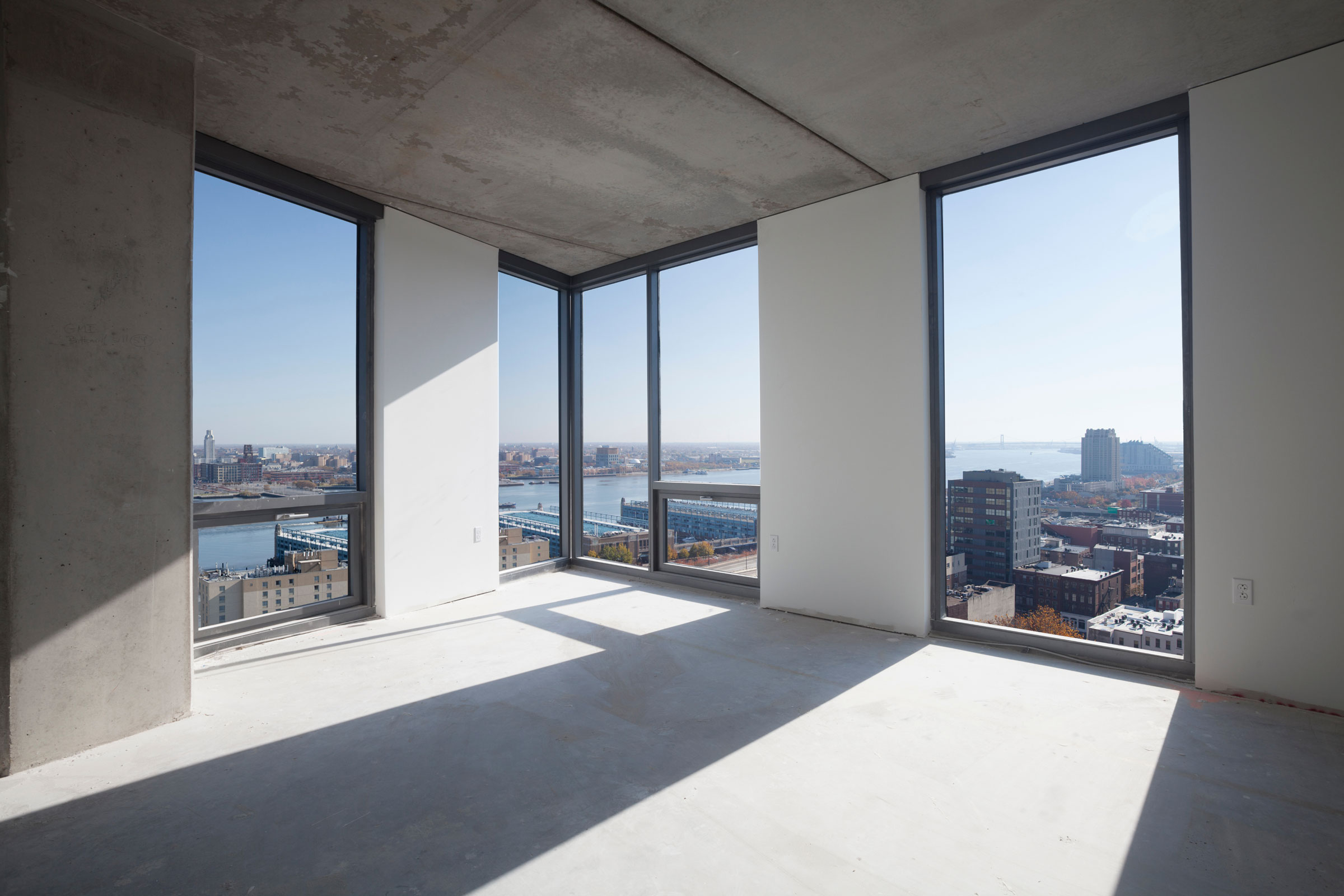 A sun-drenched, empty room with large corner windows offers panoramic views of a city skyline and river, creating a serene and spacious atmosphere.