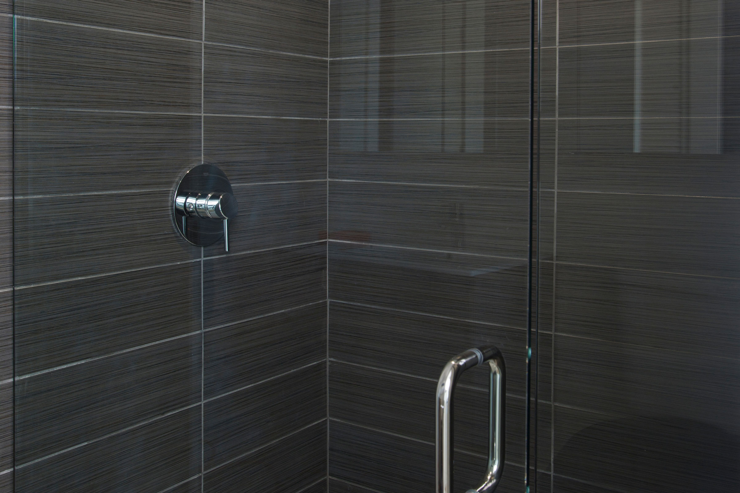 Sleek modern shower with dark gray tiles and a glass door. Chrome handle and control knob add a minimalist, elegant touch. Calm, sophisticated ambiance.