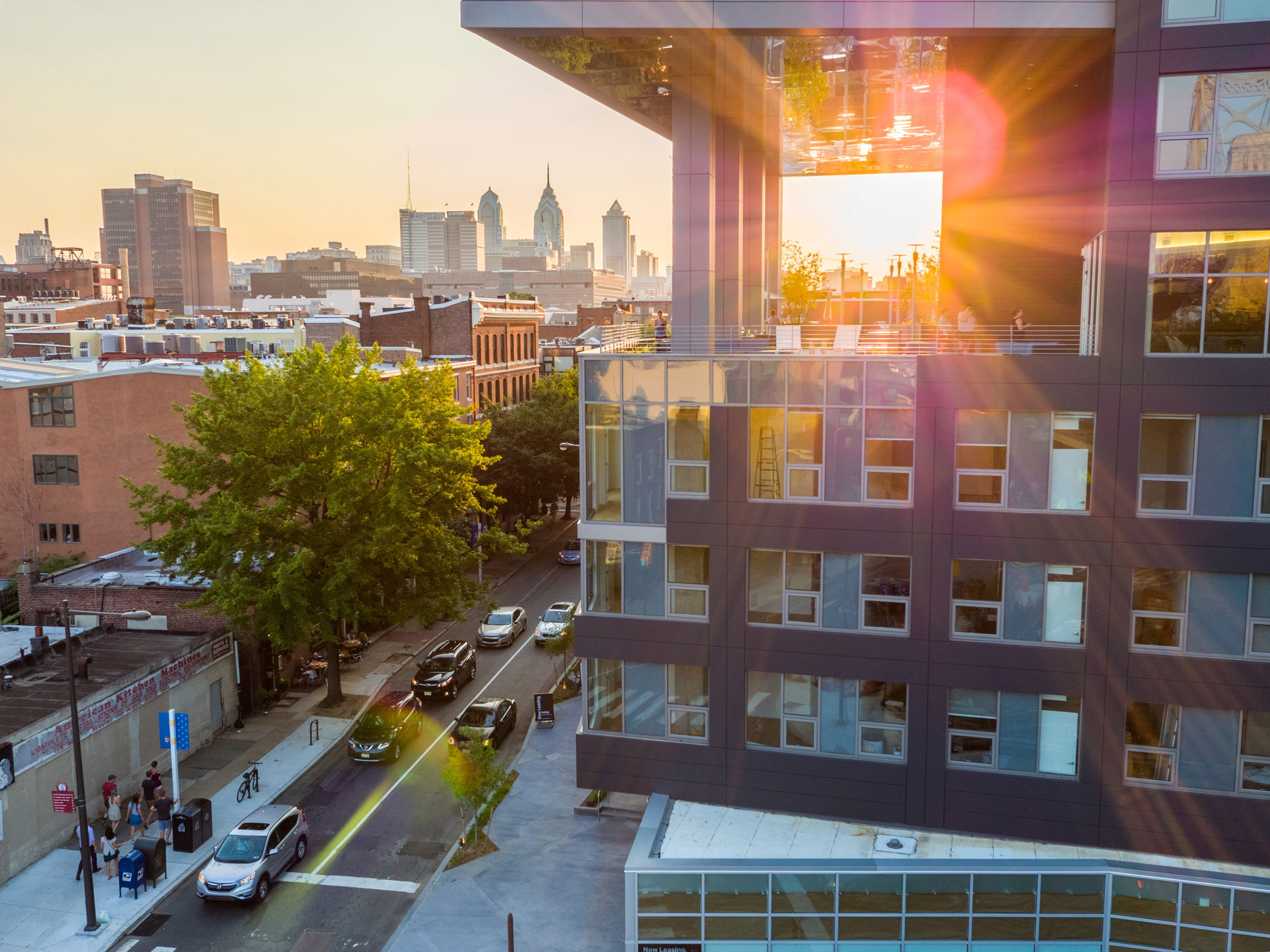 A modern building with reflective glass windows captures a vibrant sunset. Sun rays create colorful flares, while a cityscape and street activity are visible below.