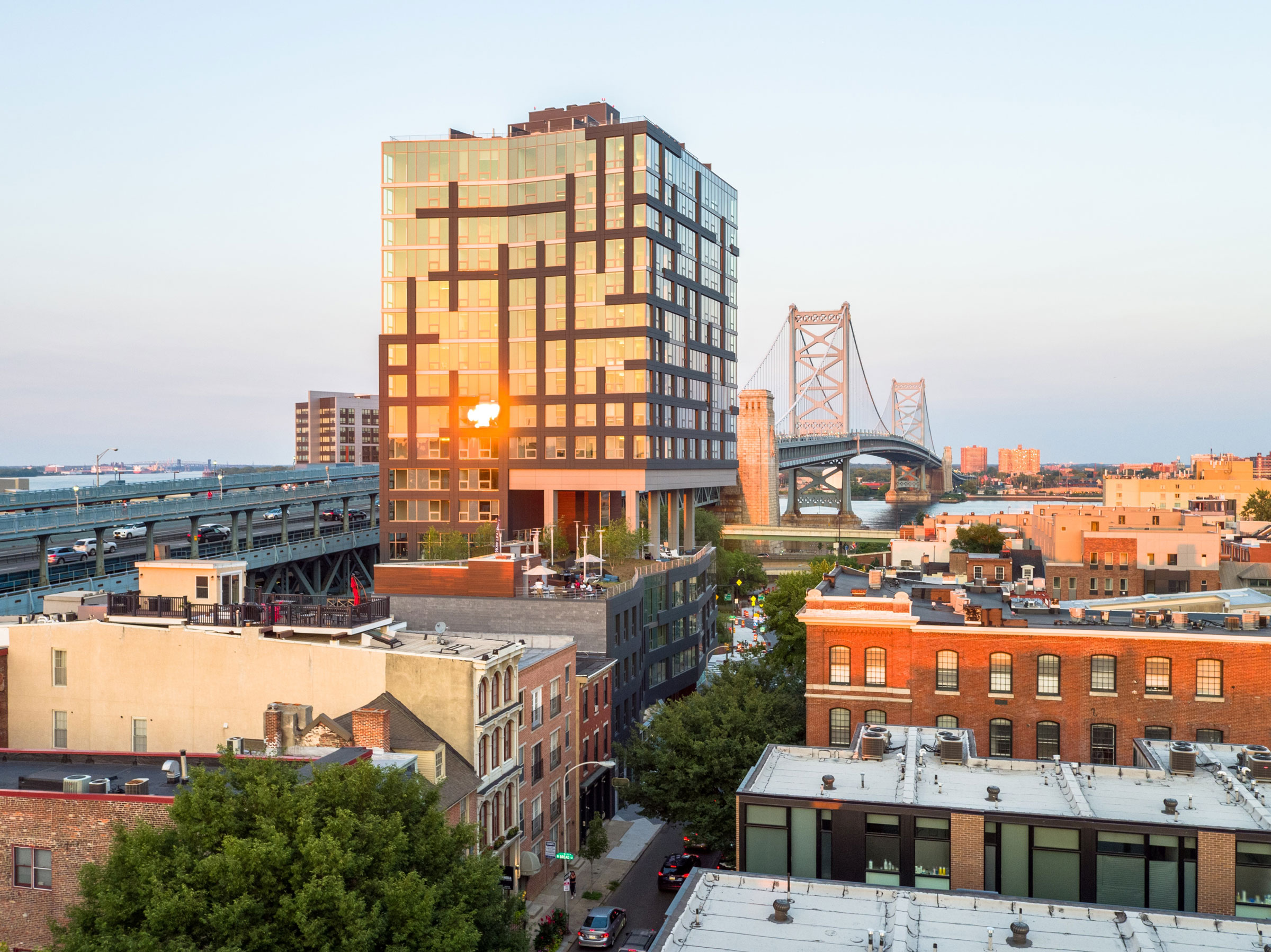 Cityscape at sunset with a modern glass building reflecting sunlight, historic brick structures, a suspension bridge, and a calm, clear sky.