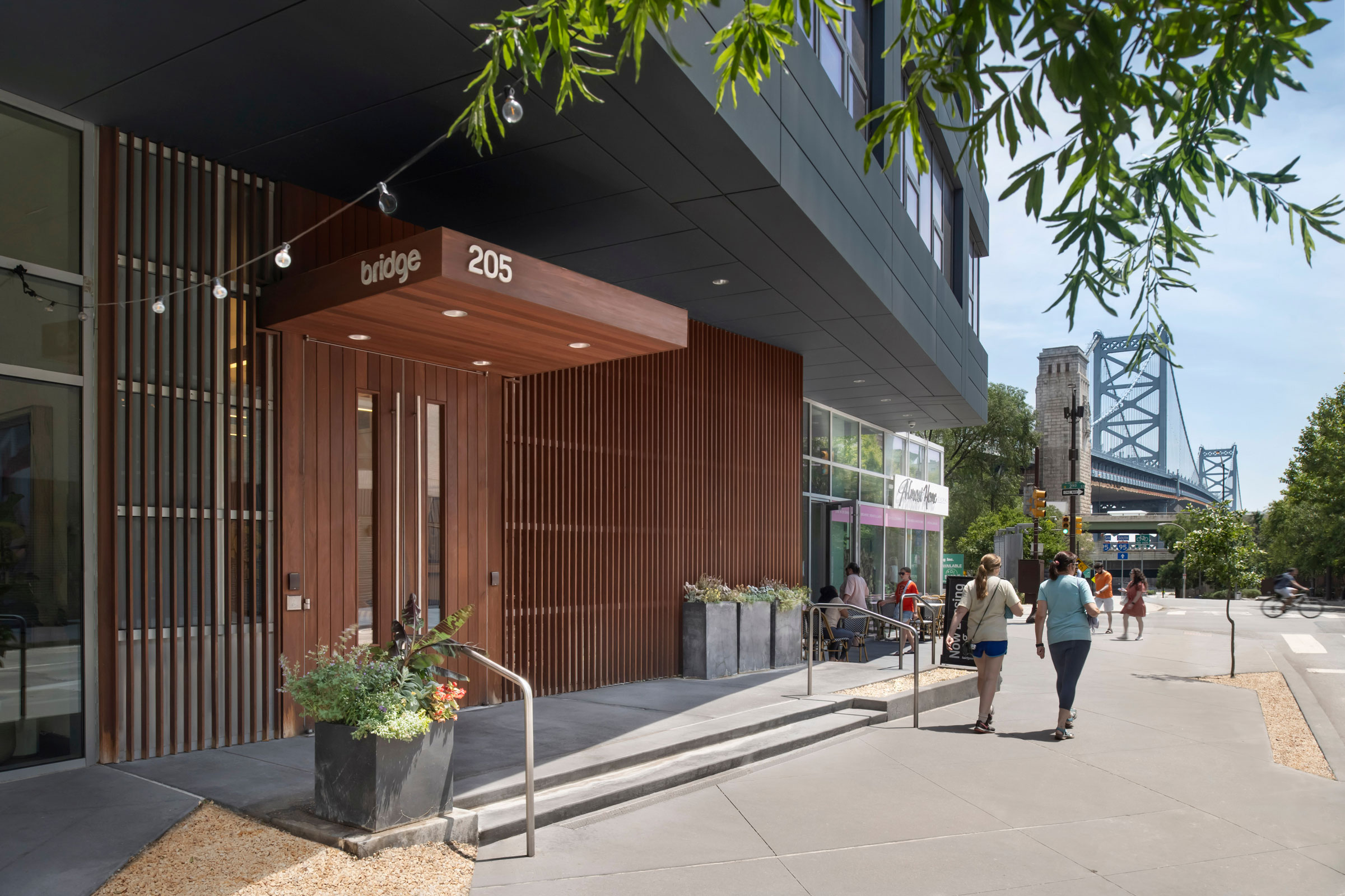 Modern urban scene with a wooden facade entrance labeled "Bridge 205" on a sunny day. People walk on a sidewalk near a bridge in the background.
