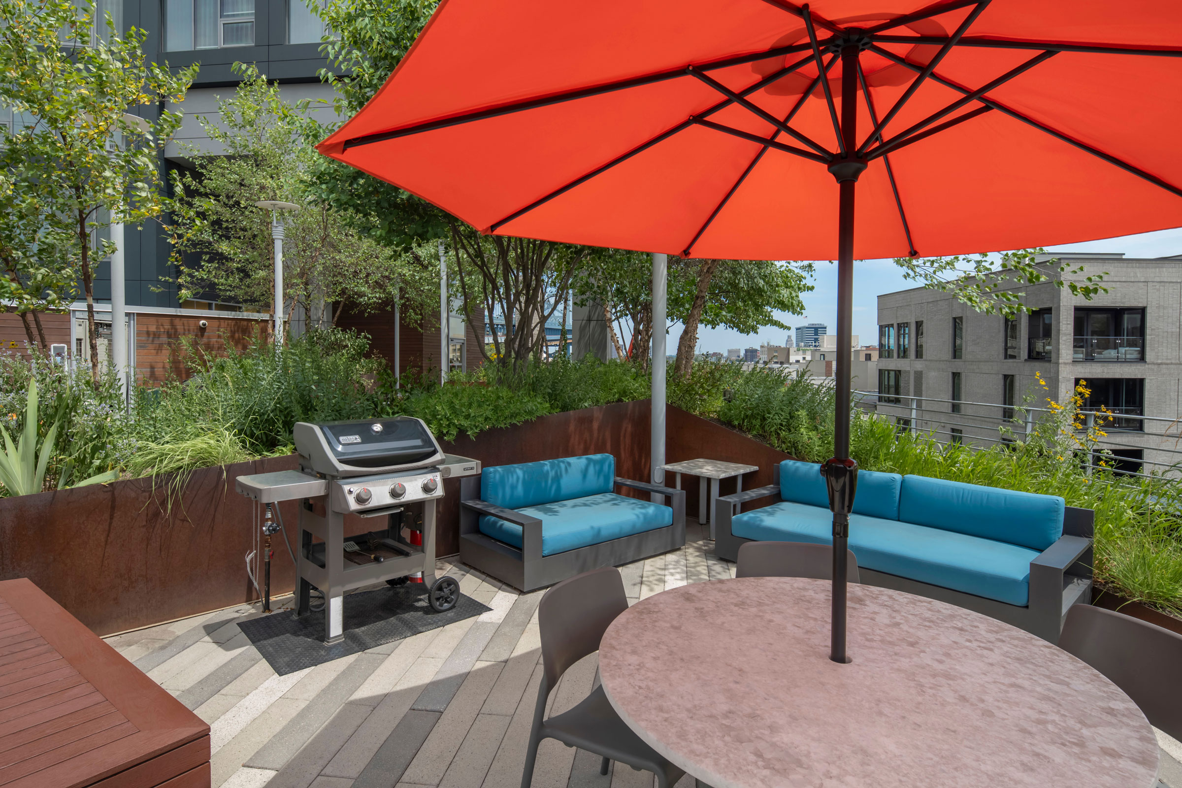Rooftop patio with a bright red umbrella, blue cushioned seating, a metal grill, and lush greenery. Modern urban buildings visible in the background.