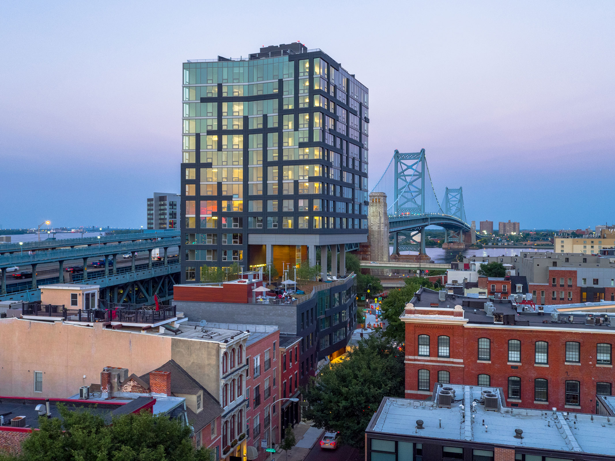 Modern glass building illuminated at dusk near a blue suspension bridge. Below are historic red-brick and pastel buildings, capturing urban contrast.