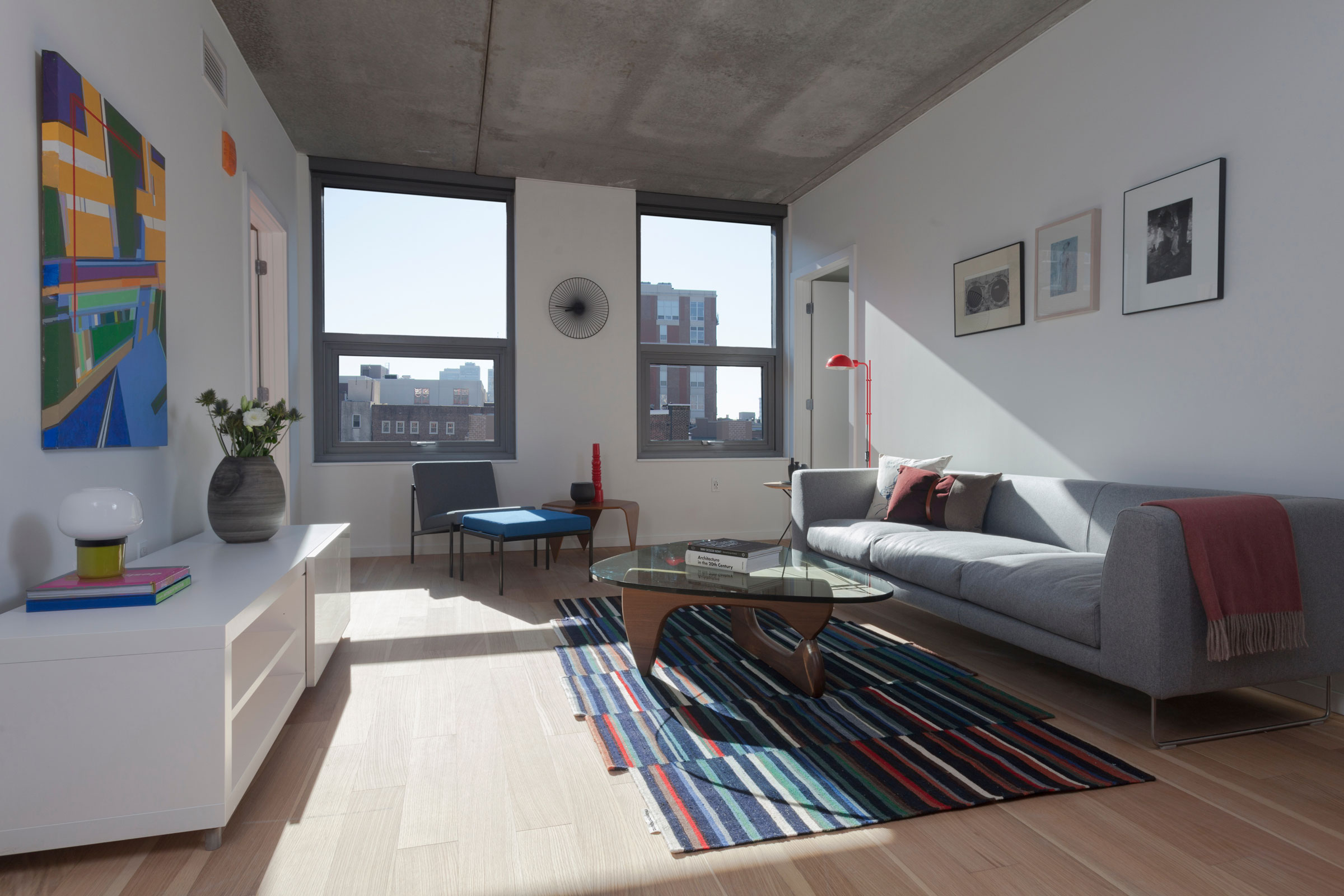 Modern living room with a grey sofa, red cushions, and a bright red lamp. A colorful abstract rug and geometric art create a vibrant, cozy feel.