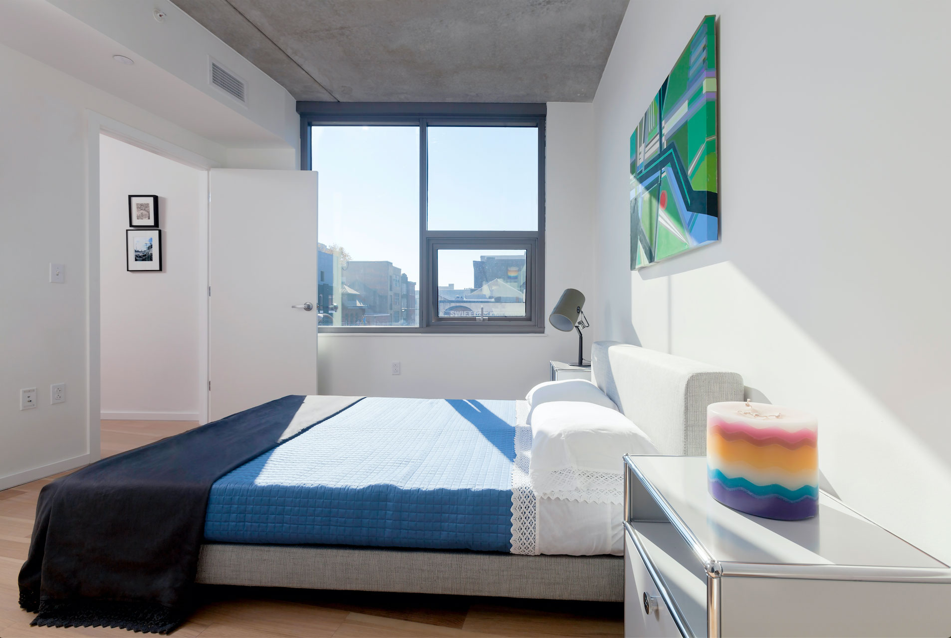 Modern bedroom with a minimalist design, featuring a bed with a blue quilt, abstract wall art, and a colorful candle on a bedside table. Bright sunlight streams through a large window, creating a calm and airy atmosphere.