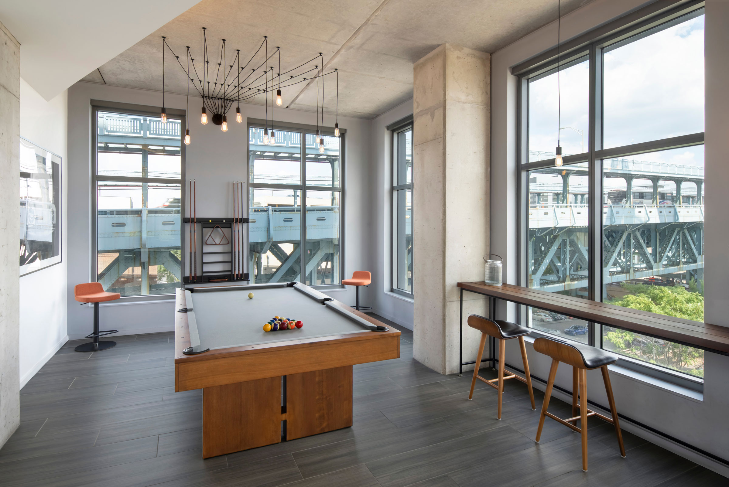 Modern game room with a wooden pool table centered under a stylish chandelier, large windows with bridge view, and bar stools for seating. Bright and airy.