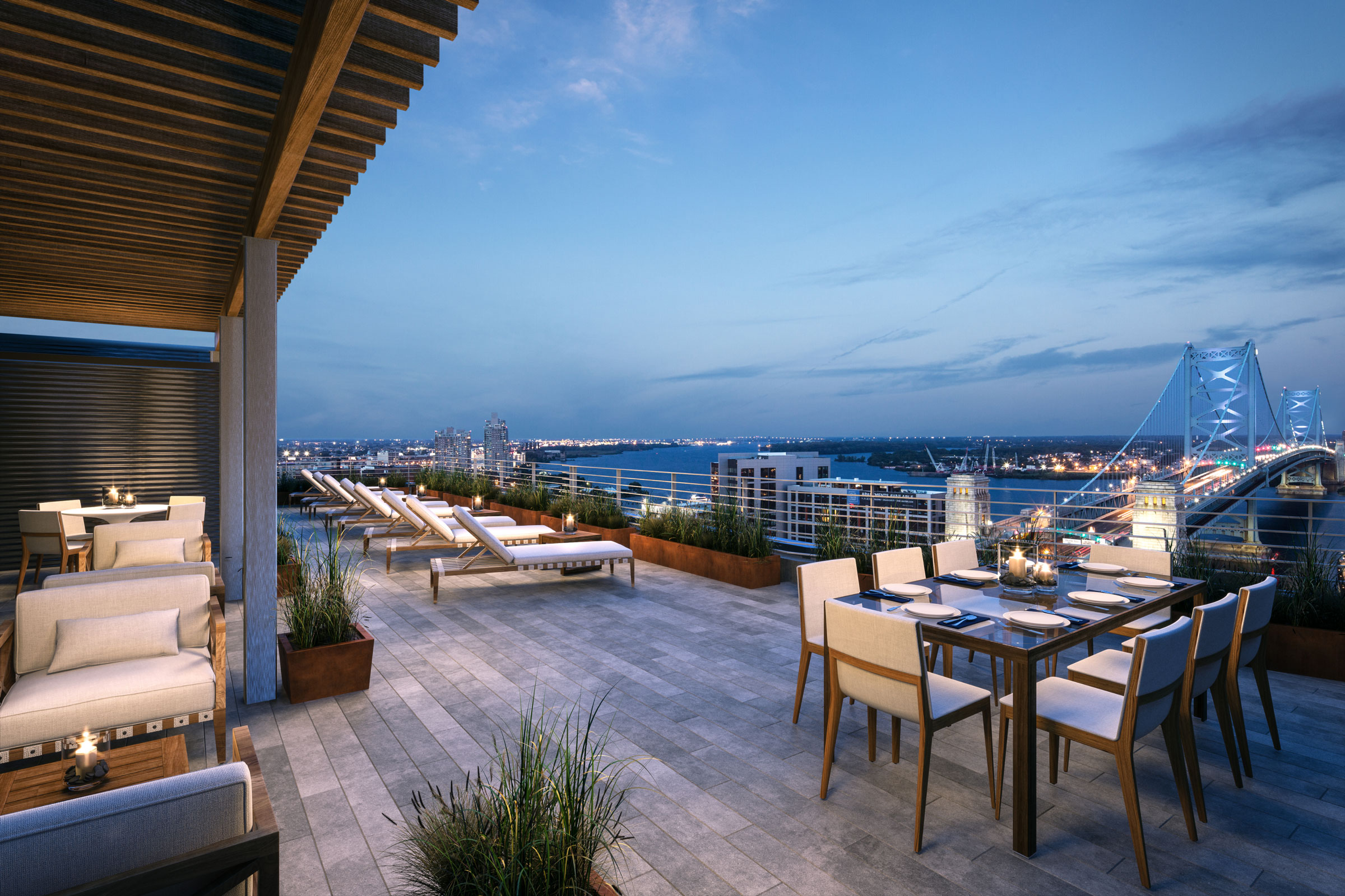 Rooftop lounge at dusk overlooking a lit bridge and cityscape. Features cozy seating, dining setup, and loungers, creating a relaxed, elegant ambiance.