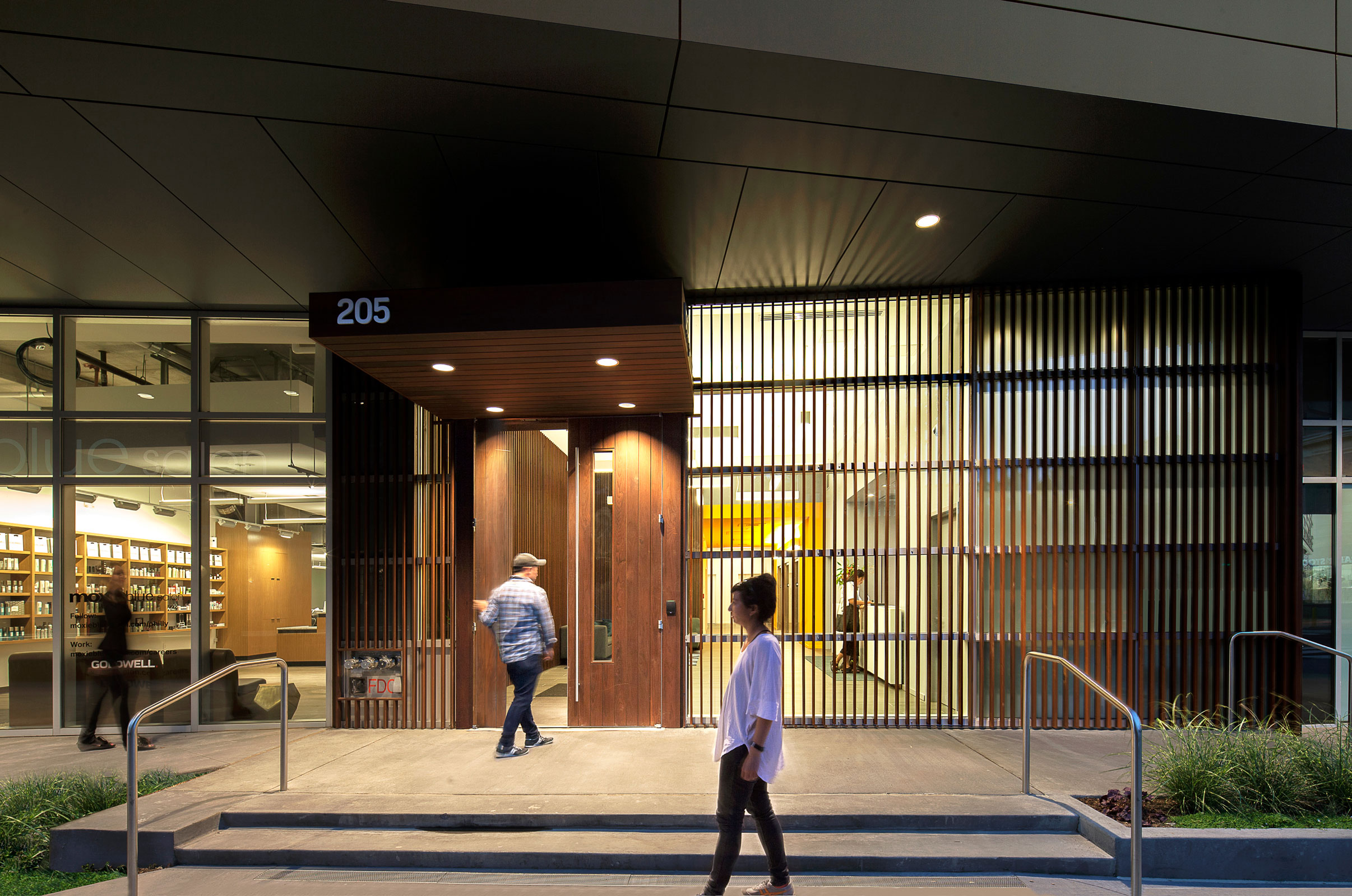Modern building entrance at dusk, warmly lit. A person in a checkered shirt enters through wood-paneled doors, while another walks on the sidewalk.