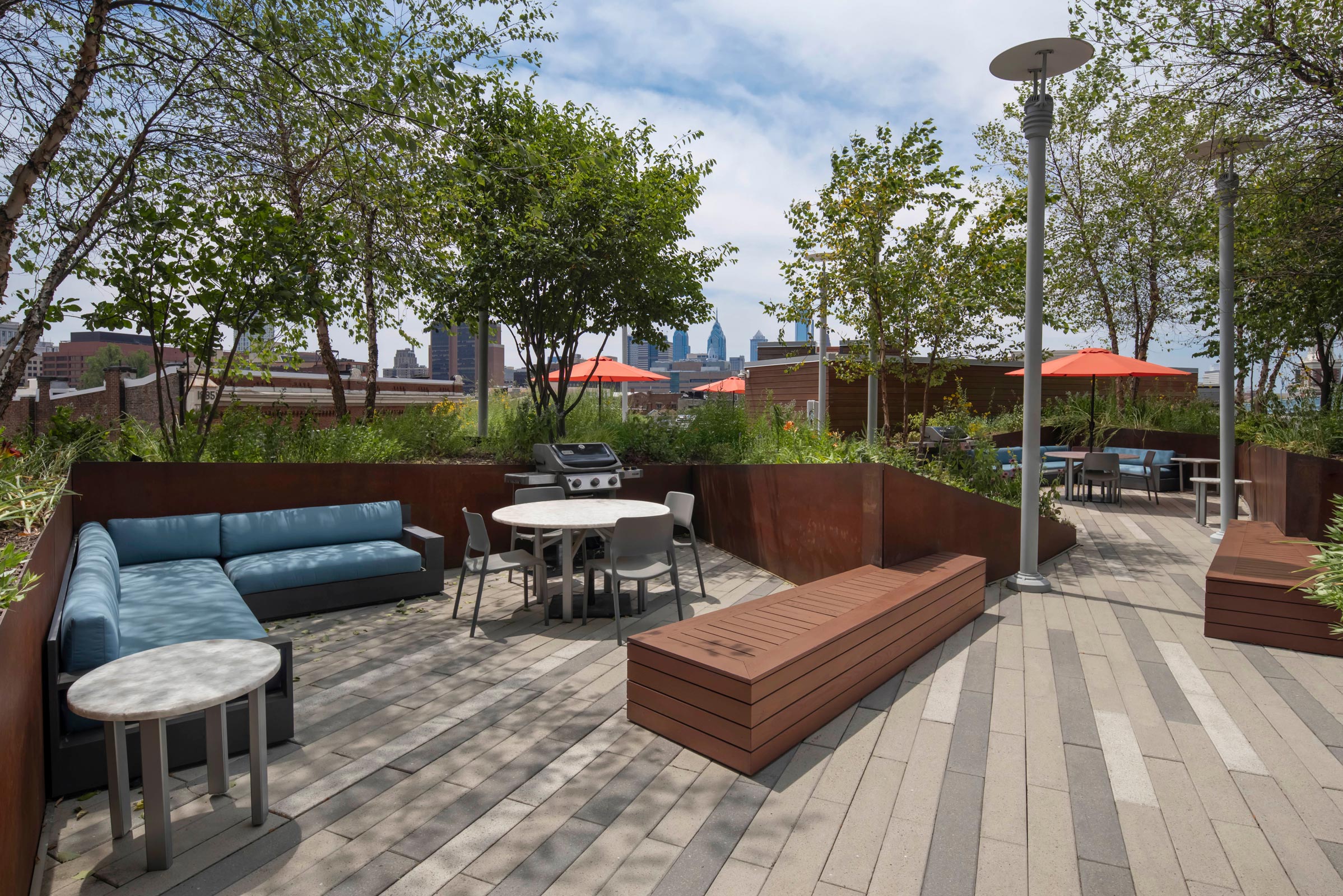 Rooftop patio with modern outdoor seating, a round table, and a grill. Surrounded by trees and city skyline in the background under a clear sky.