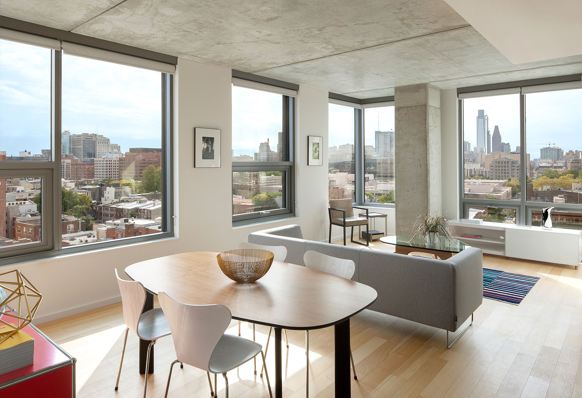 Modern apartment interior with large windows showcasing a cityscape. Features a wooden dining table, gray sofa, and light wood flooring. Bright and airy atmosphere.