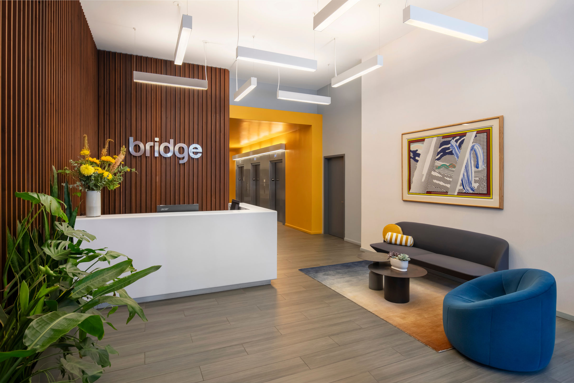A modern reception area with a sleek white desk, wooden slat wall, and "Bridge" logo. Bright yellow hallway, stylish seating, vibrant artwork, and plants create a welcoming vibe.
