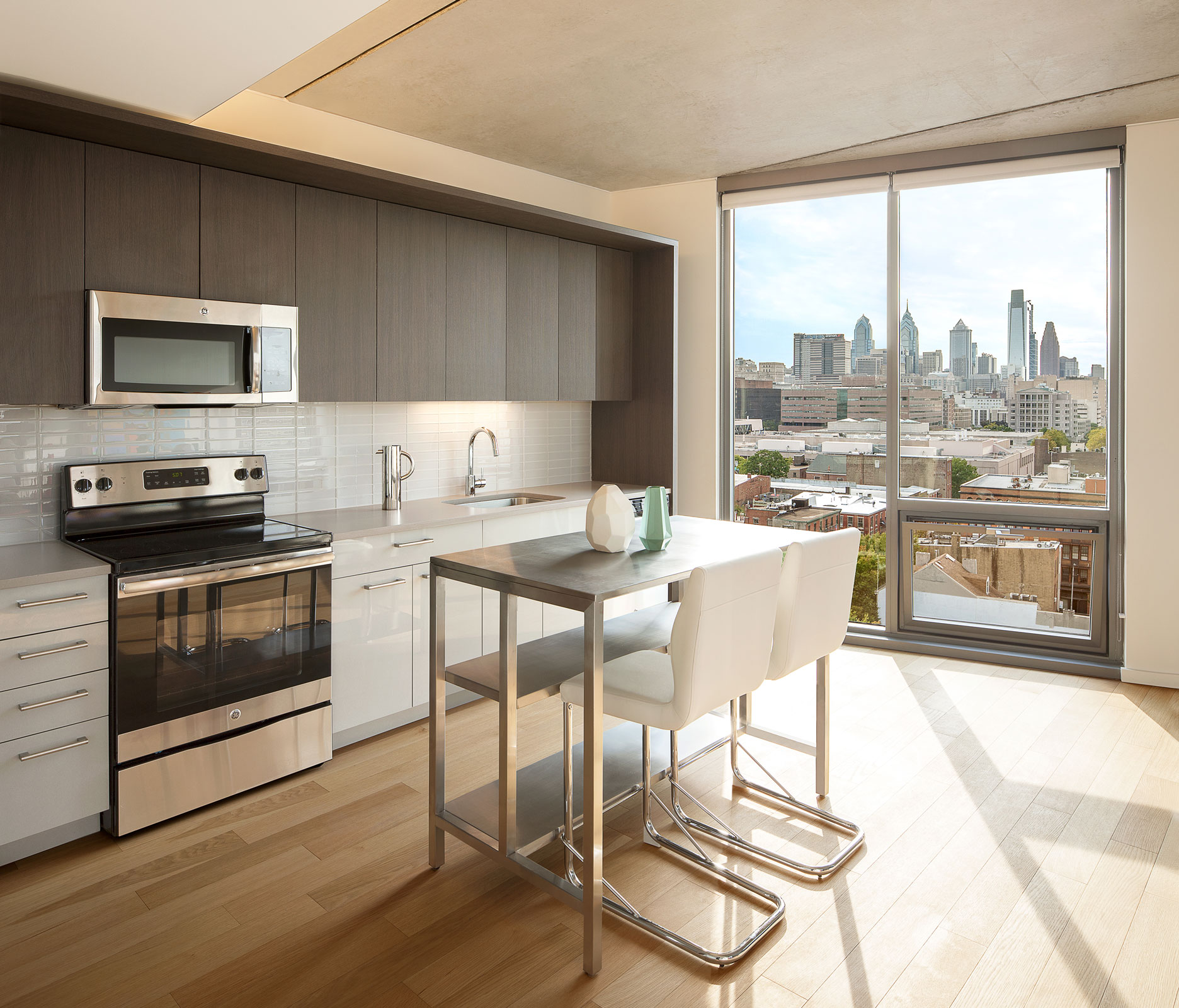 Modern kitchen with dark wood cabinets, stainless steel appliances, and a sleek island with white chairs. Large window offers a city skyline view.