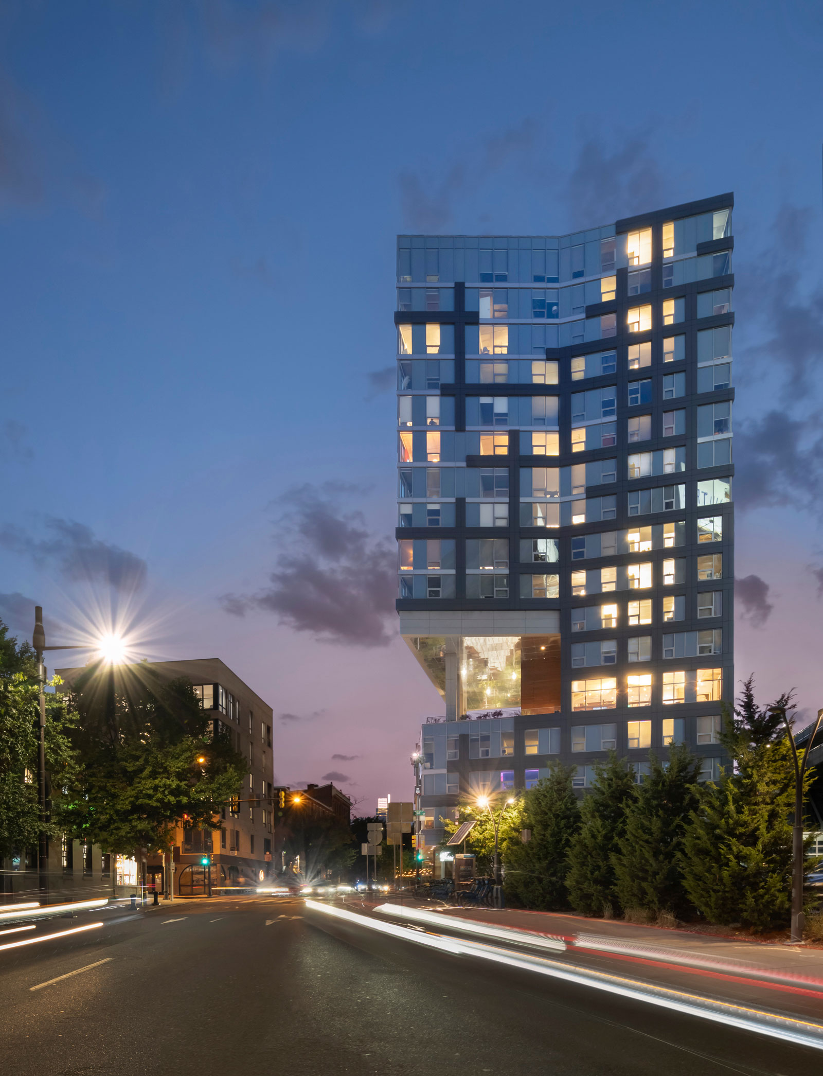 Modern building illuminated at dusk, featuring a geometric design with large windows. Streetlights and blurred car lights create a dynamic cityscape.