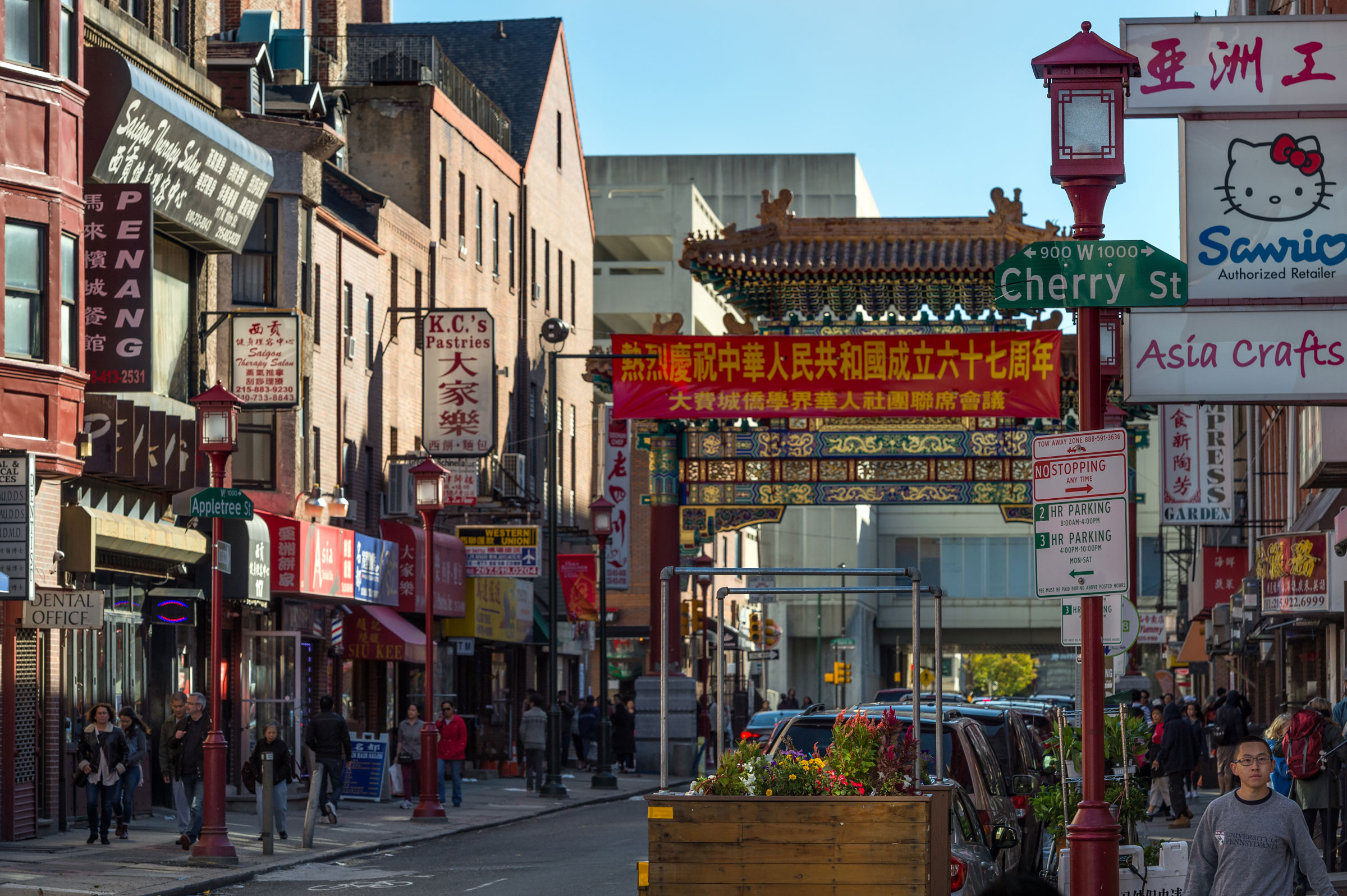 Bustling Chinatown street with colorful signs in Chinese characters, a decorative archway, and pedestrians walking under sunny skies in an urban setting.