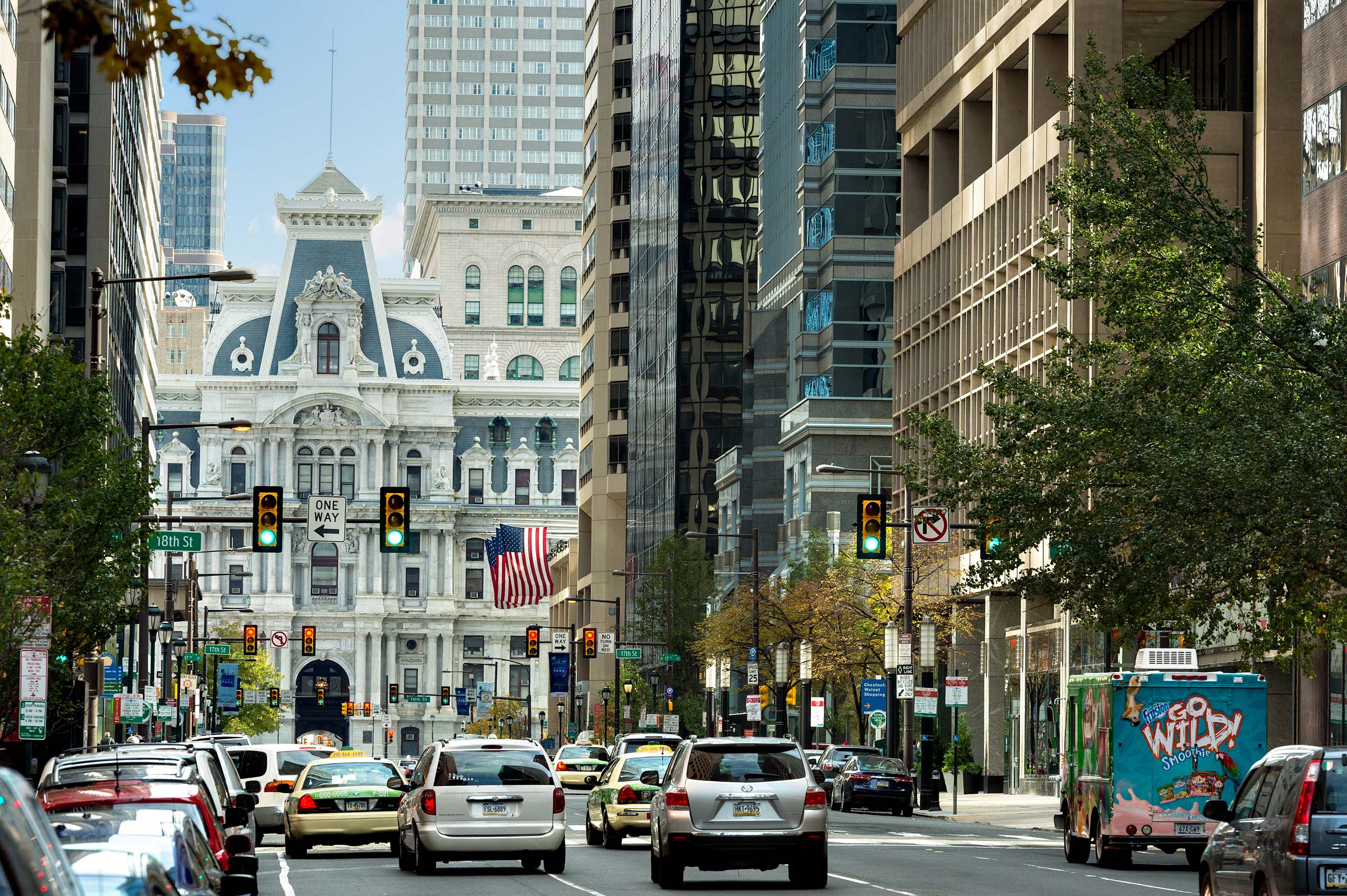 Bustling city street with cars and taxis, flanked by tall buildings. A historic building with ornate architecture is centered, flying a large American flag.