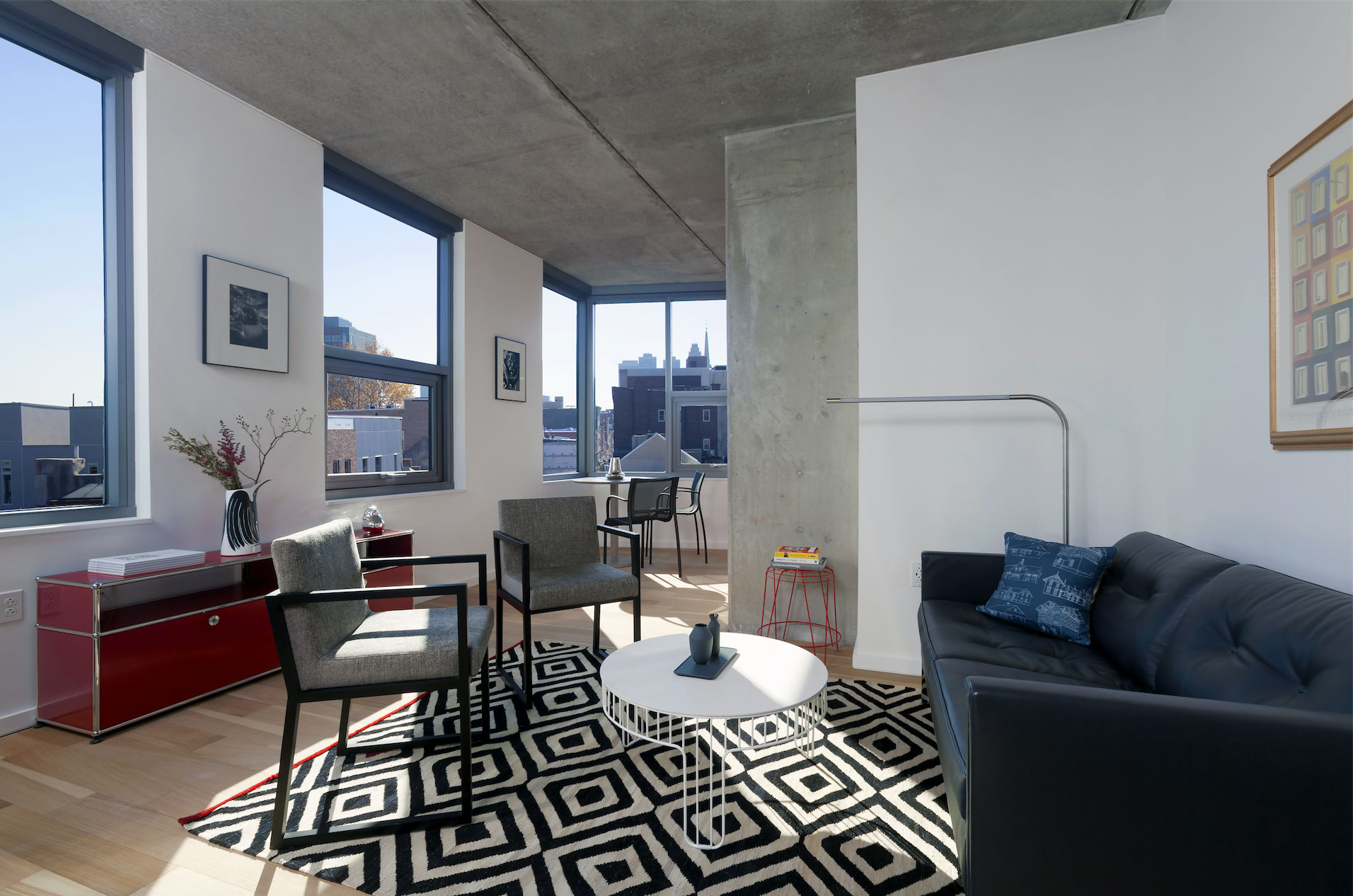Modern living room with black leather sofa, geometric rug, and minimalist furniture. Large windows with cityscape views, creating an airy feel.