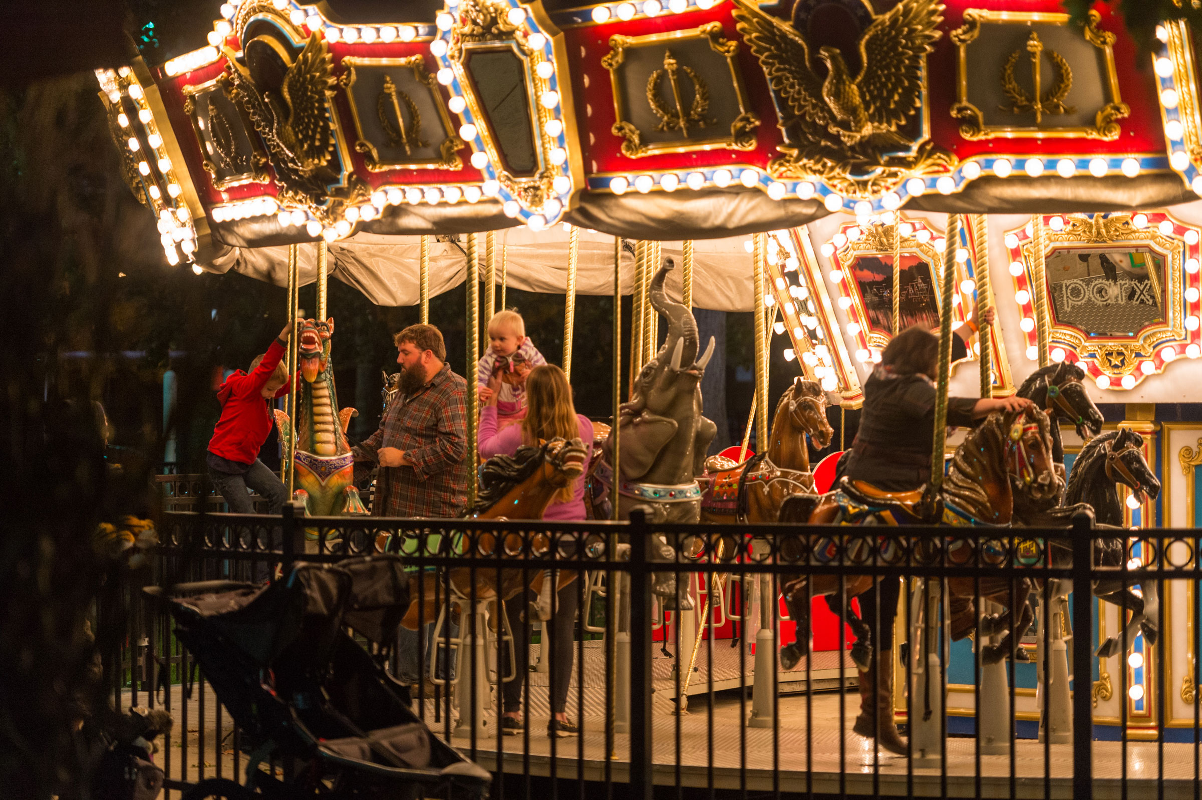 A brightly lit carousel with children riding colorful horses at night. A man in a plaid shirt holds a baby, surrounded by a joyful atmosphere.