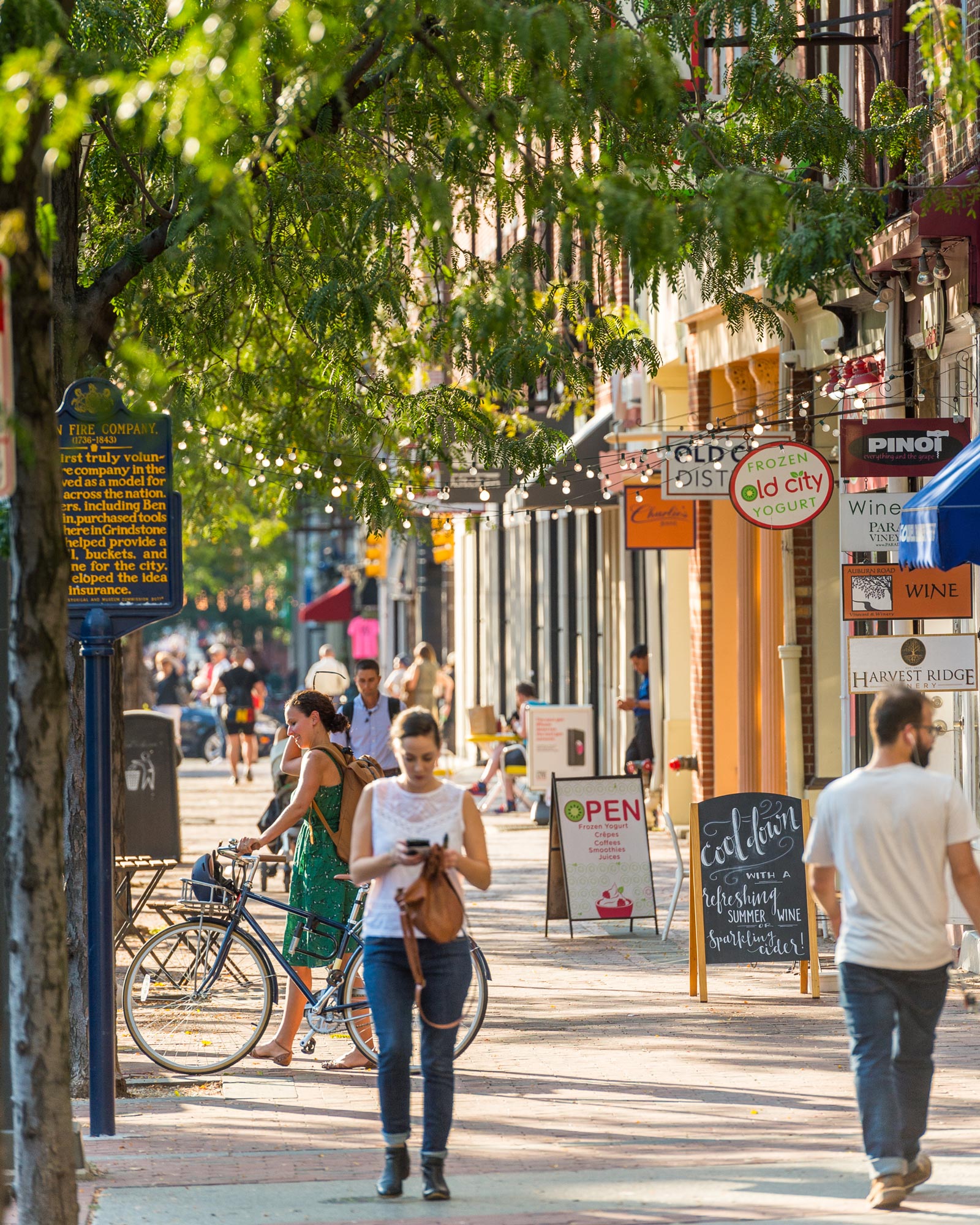 A lively street scene in a sunlit city area. People walk and cycle by shops with bright signs and leafy trees overhead, creating a vibrant, bustling atmosphere.