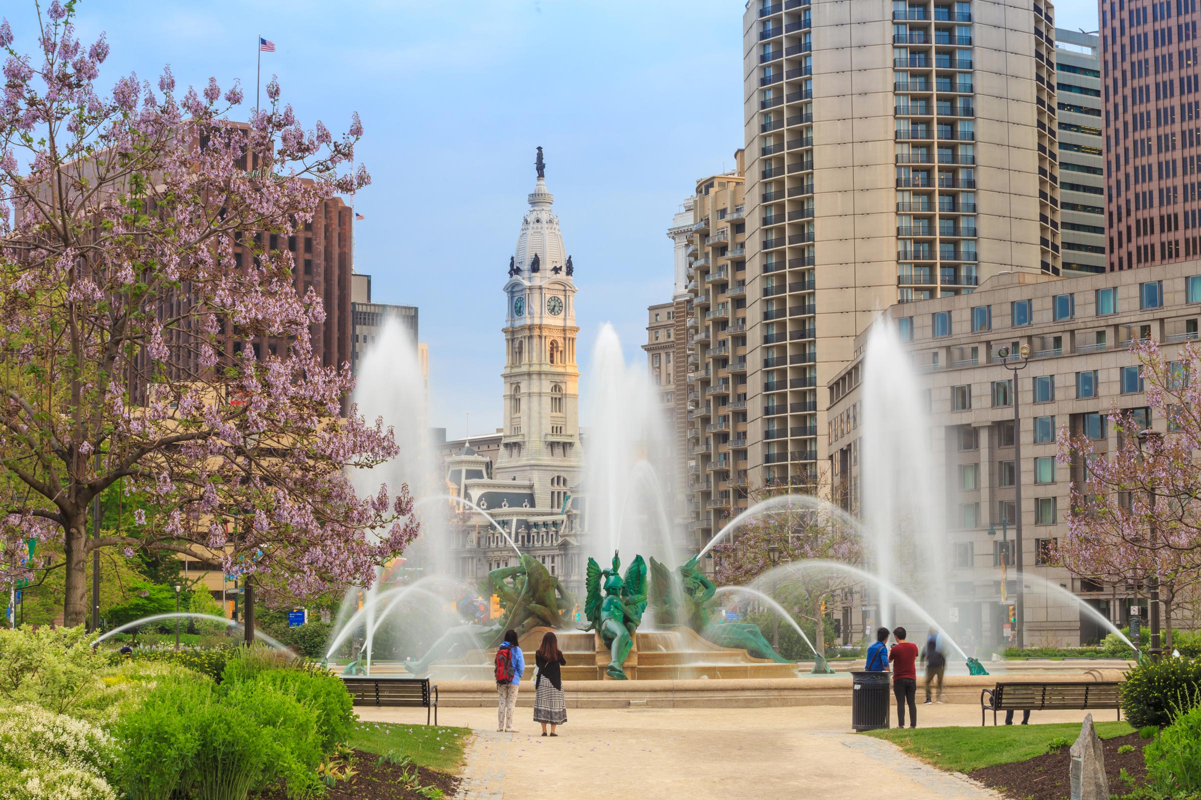 Springtime scene of a park with a large fountain and blooming trees, set against a cityscape of tall buildings and a clock tower under a blue sky.