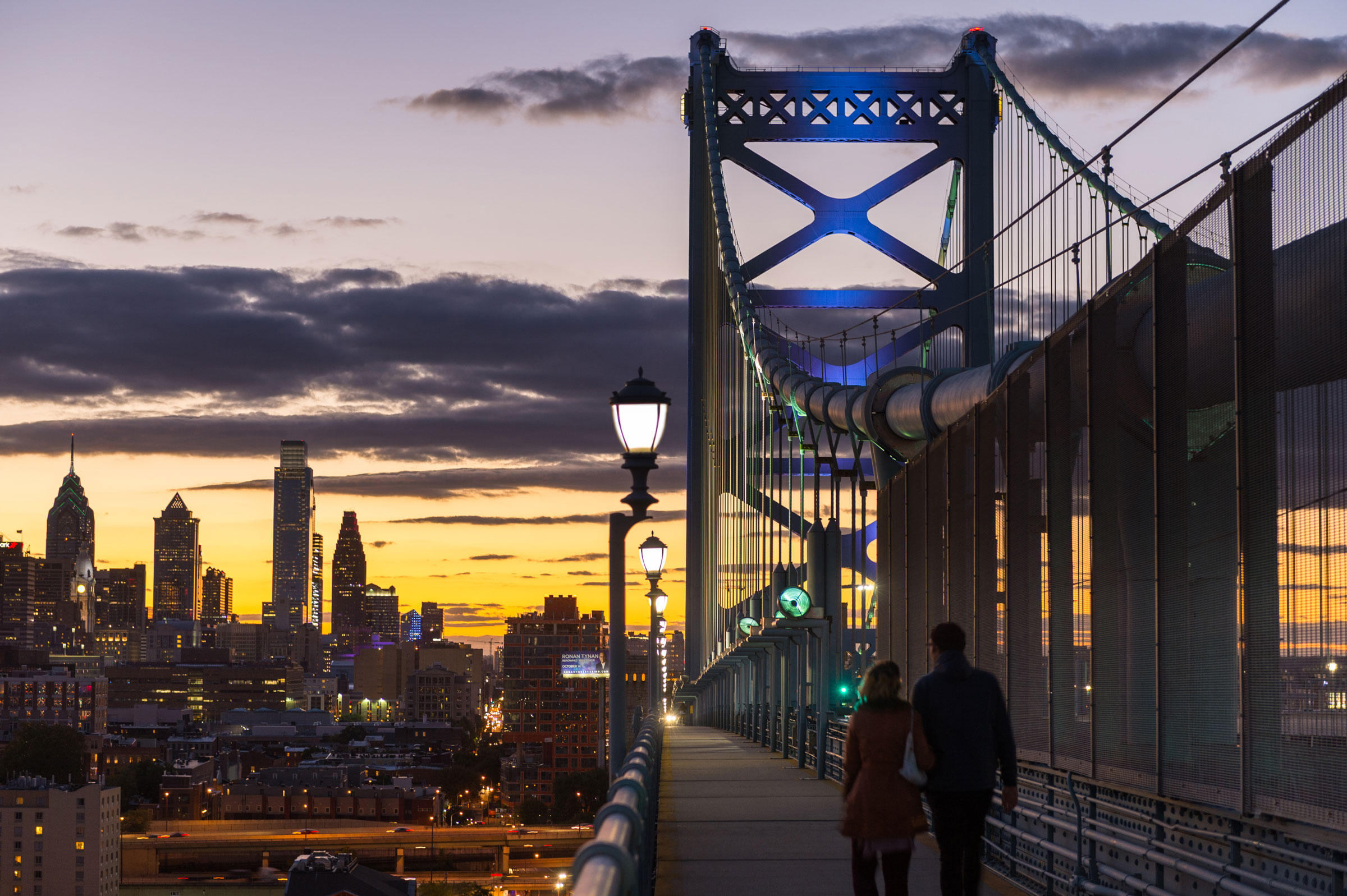 Couple walks on a bridge at sunset, silhouetted against a vibrant sky. City skyline with illuminated skyscrapers in the background. Calm, romantic atmosphere.