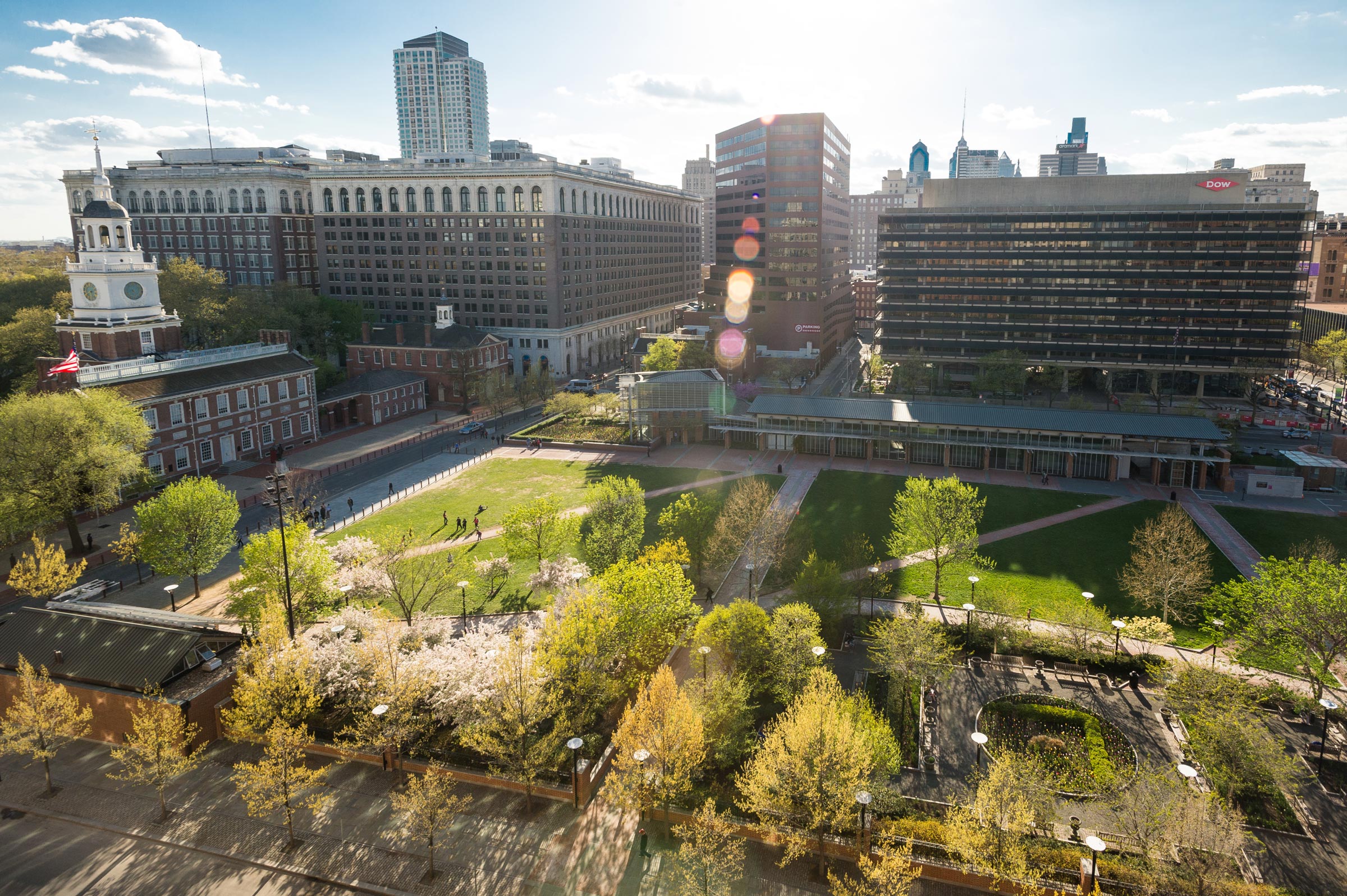 Aerial view of Independence Hall and its surrounding green park in Philadelphia. Sunlight bathes the scene, creating a bright, lively atmosphere.