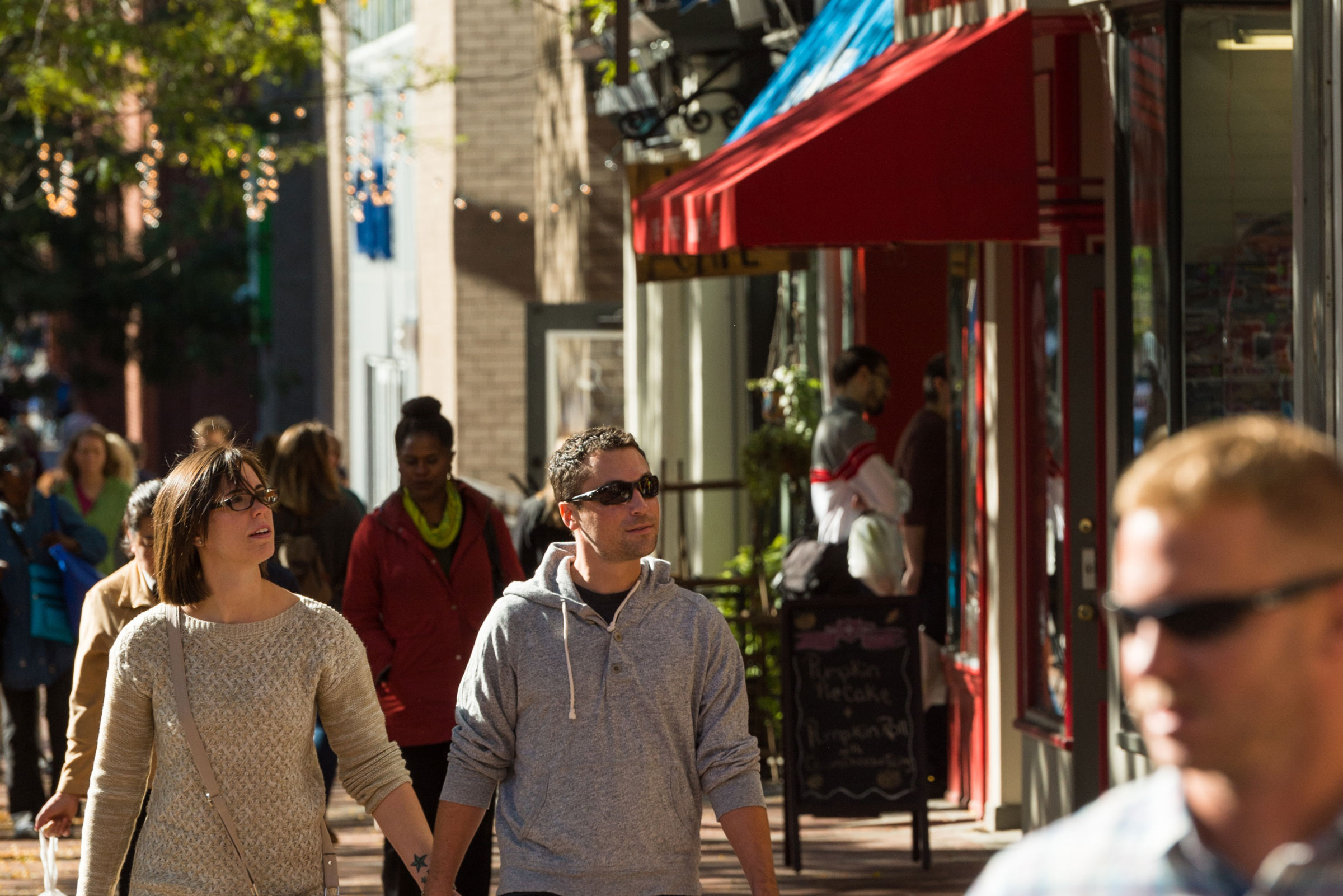 A couple walks hand in hand down a lively urban street, framed by a red canopy and string lights. The scene conveys a relaxed, sunny day.