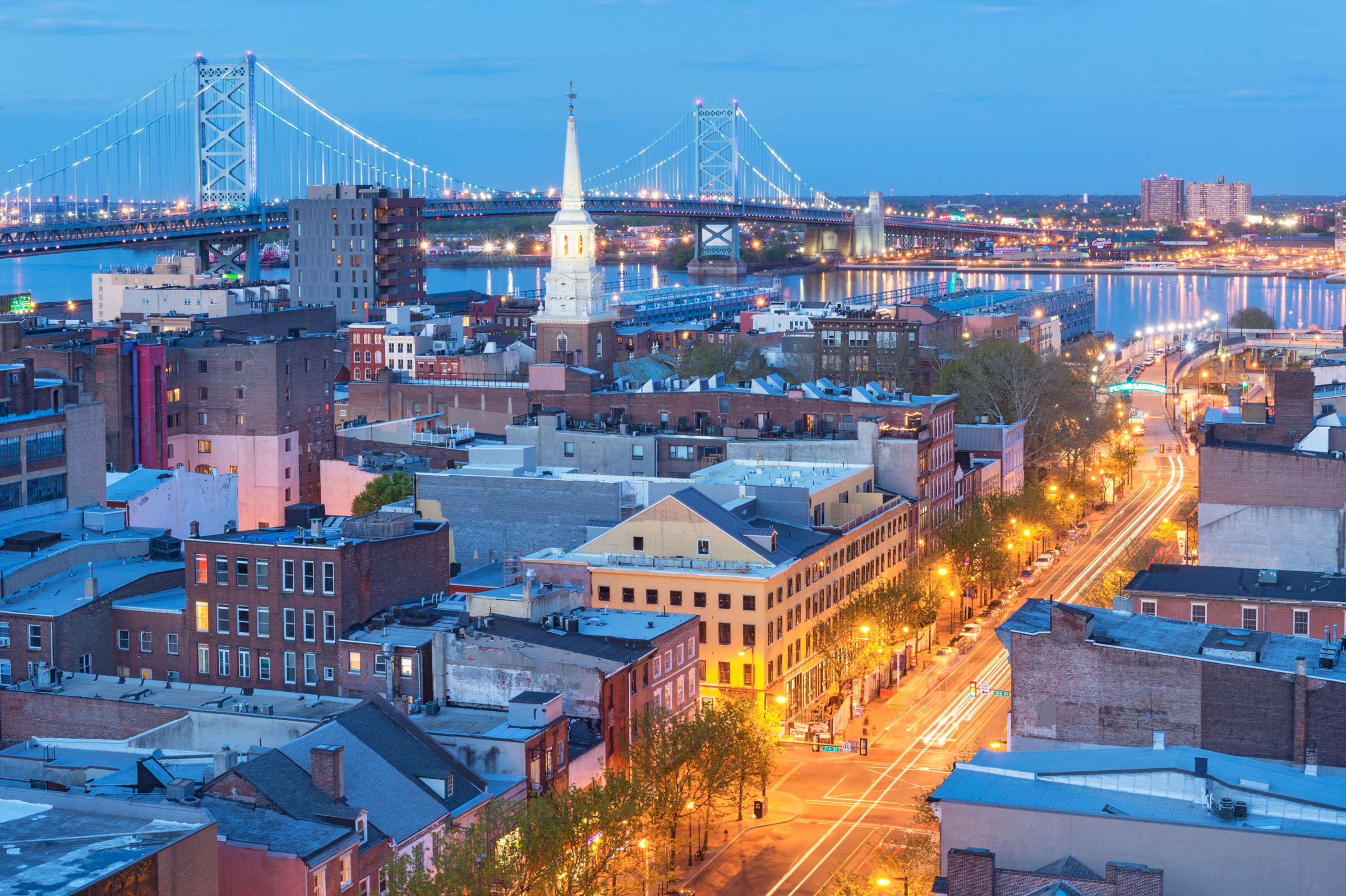 Cityscape at dusk with a lit bridge over a river, a church steeple, and glowing streetlights on a bustling street, conveying urban vibrancy.