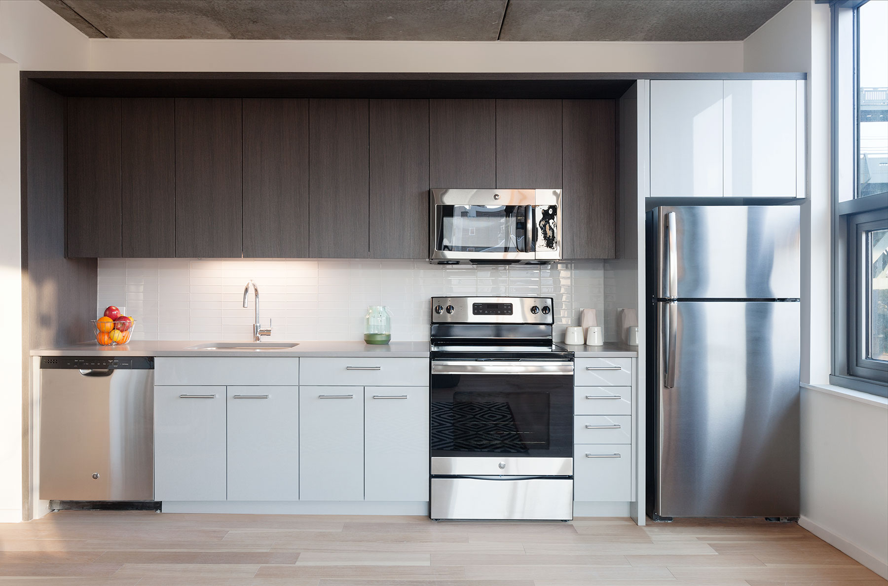 Modern kitchen with sleek gray cabinets, stainless steel appliances, and a fruit bowl. Sunlight streams through a large window, creating a cozy atmosphere.