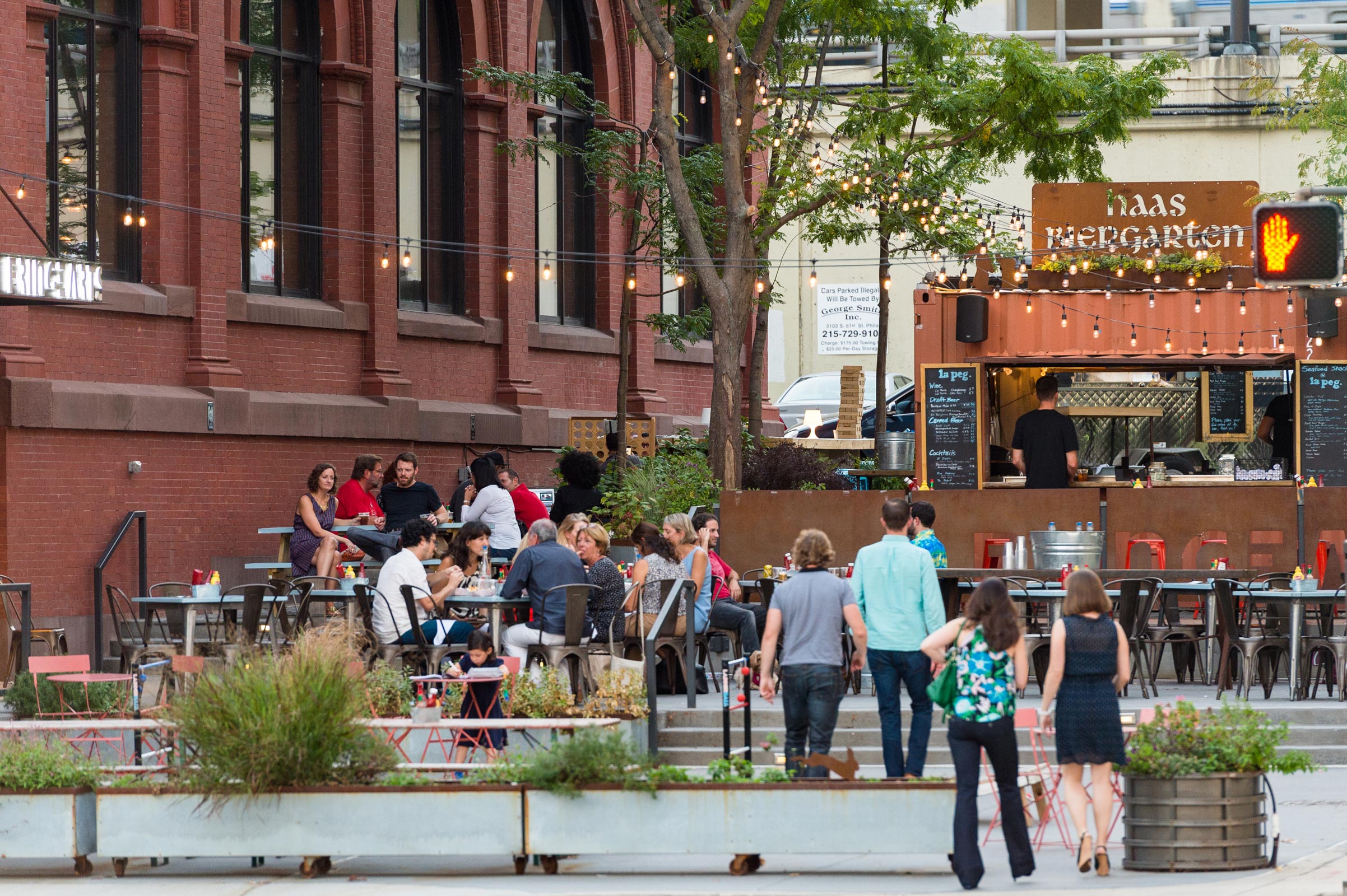 Outdoor seating area beside a brick building, filled with people dining. A food stand labeled 'Haas Biergarten' under string lights creates a lively atmosphere.