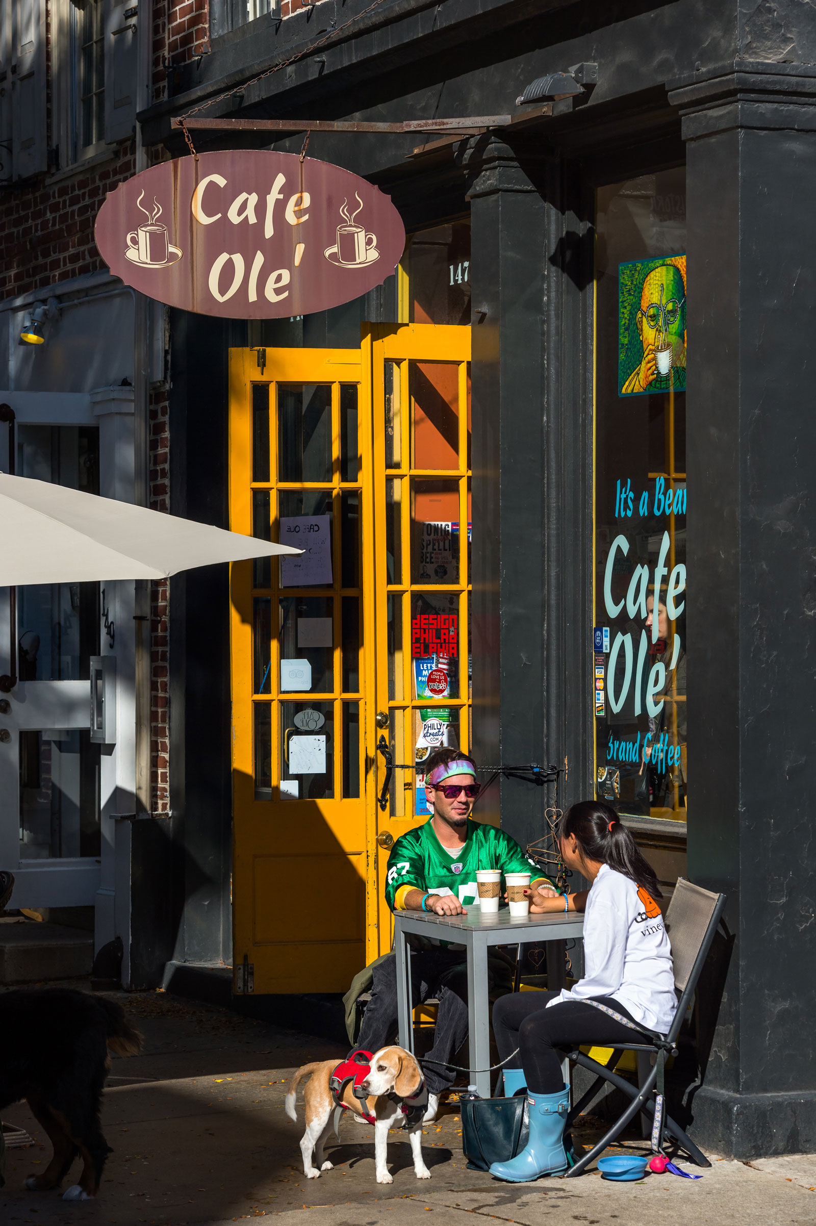 A couple sits outside Café Olé, enjoying coffee on a sunny day. A beagle on a leash stands nearby, and a vibrant yellow door adds warmth.