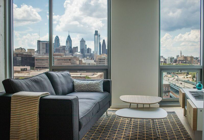 Modern living room with a dark gray sofa, patterned rug, and nested coffee tables. Large windows show a city skyline under partly cloudy skies.