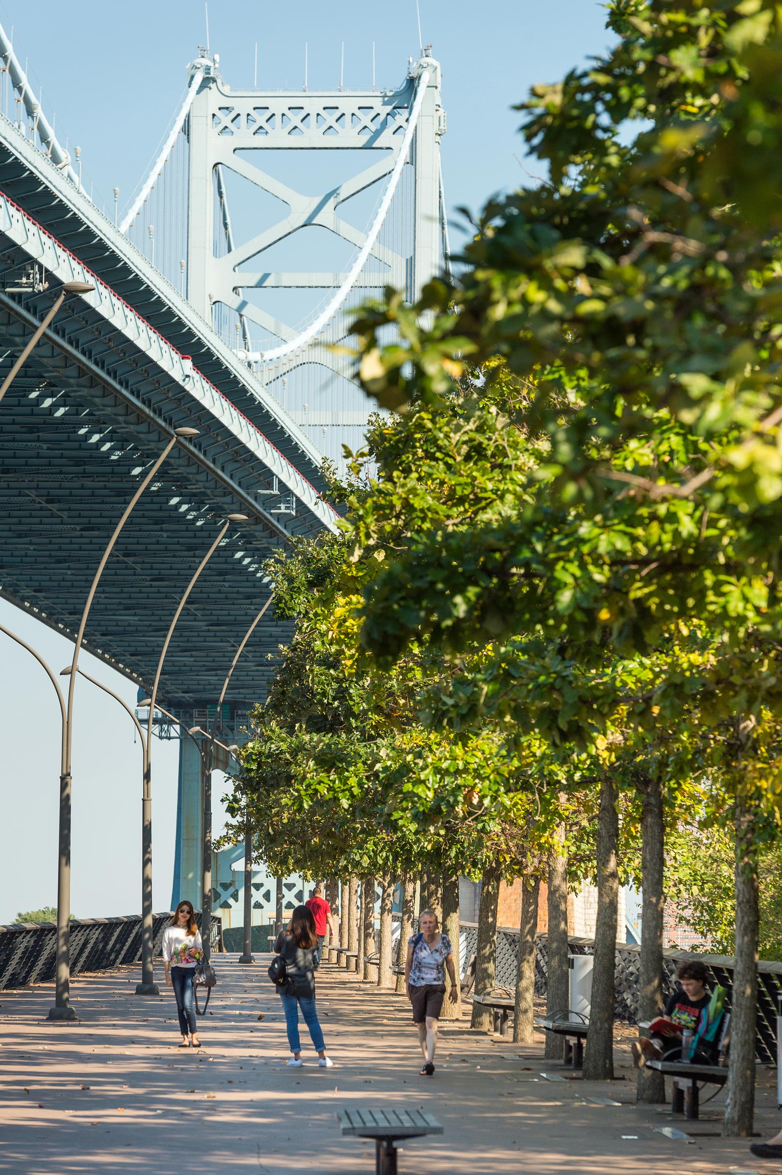 People stroll along a tree-lined walkway beside a large suspension bridge on a sunny day. The scene is calm and vibrant, evoking a sense of leisure.