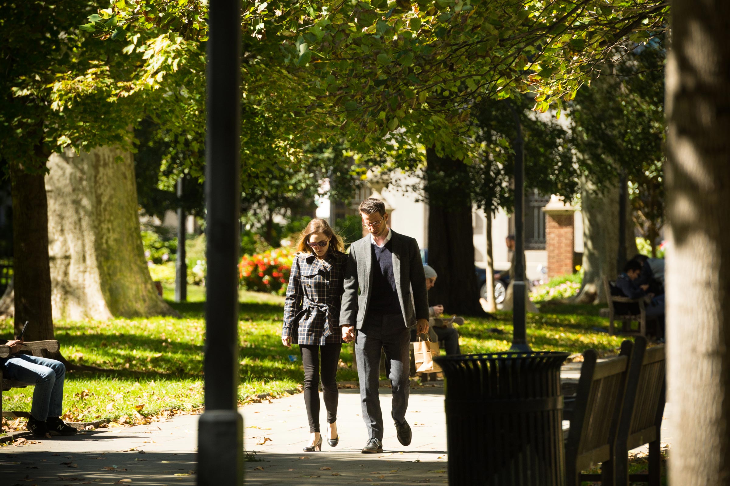 A couple walks hand in hand down a sunlit path in a park, surrounded by green trees. People sit on benches in the background, adding a peaceful ambiance.