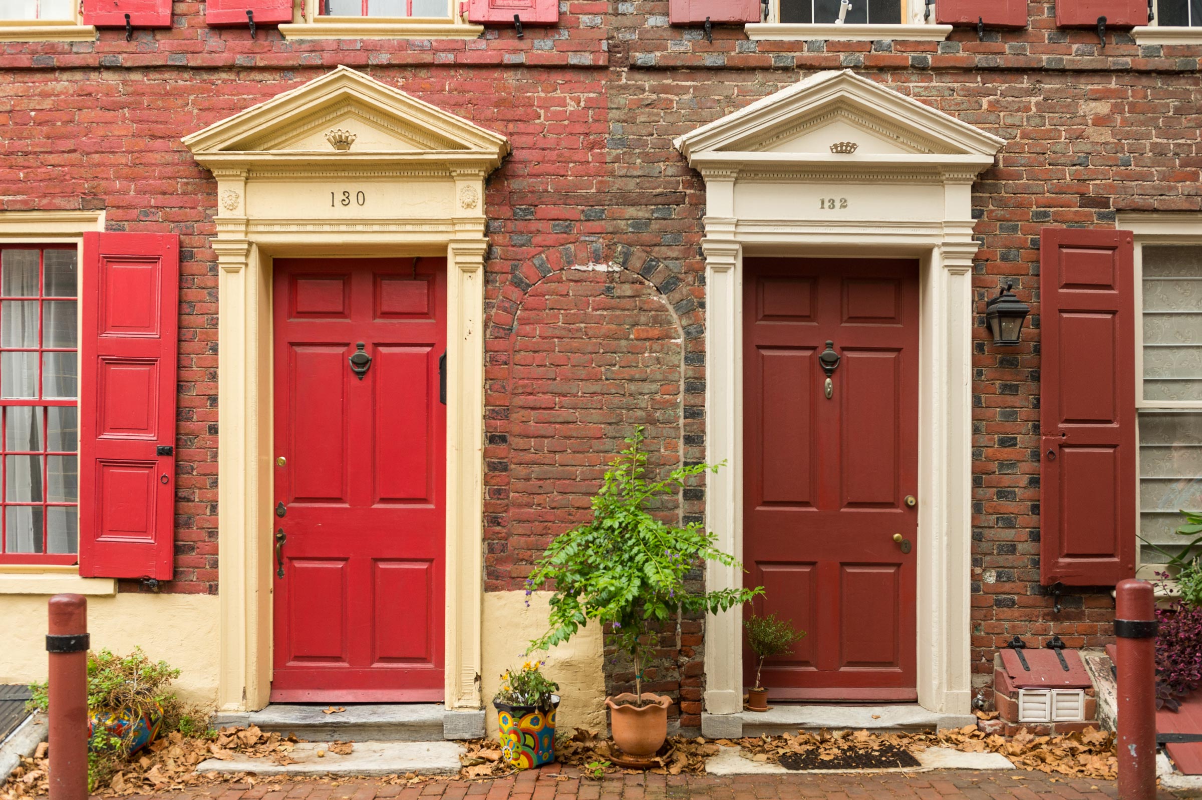 Two adjacent red doors on a brick building; door 130 with tan trim, door 132 with white trim. Potted plants and fallen leaves decorate the entrance.