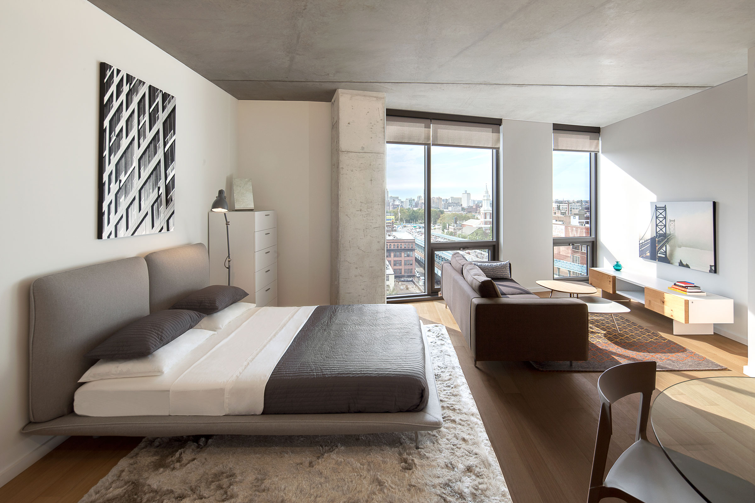 Modern bedroom with large windows and city view. Features a gray bed, abstract art, cozy rug, brown sofa, and minimal decor, conveying a calm ambiance.