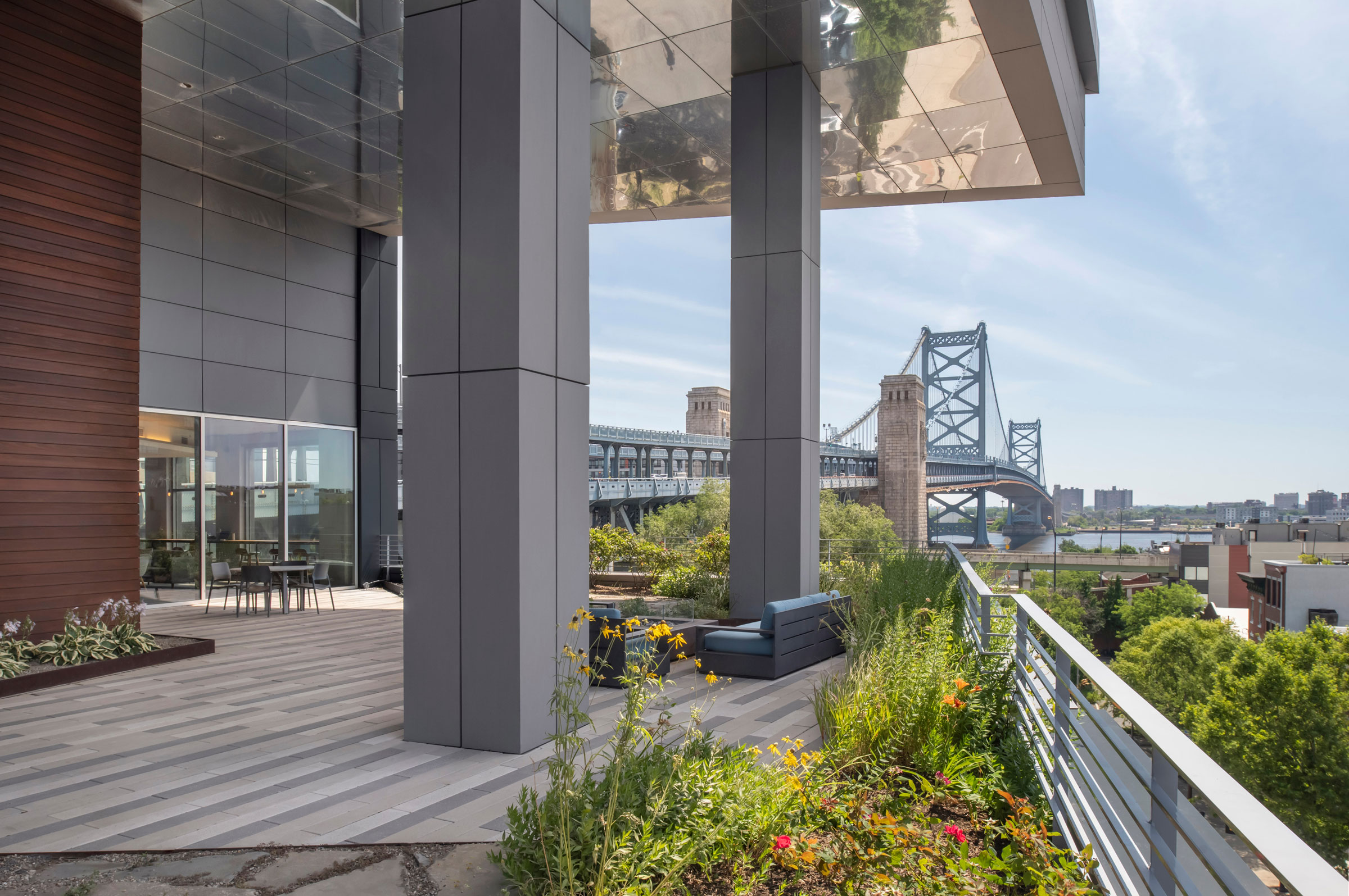 Modern terrace with gray pillars and wooden wall, overlooking a river and bridge. Flower garden on the edge, bright and serene atmosphere.