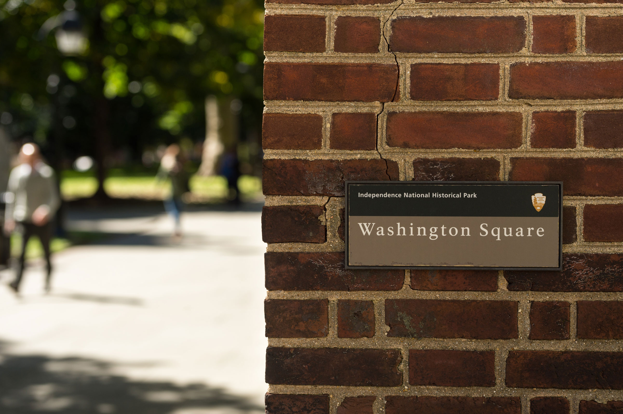 A brick wall with a sign that reads "Washington Square, Independence National Historical Park." Blurred background shows people walking and green trees.